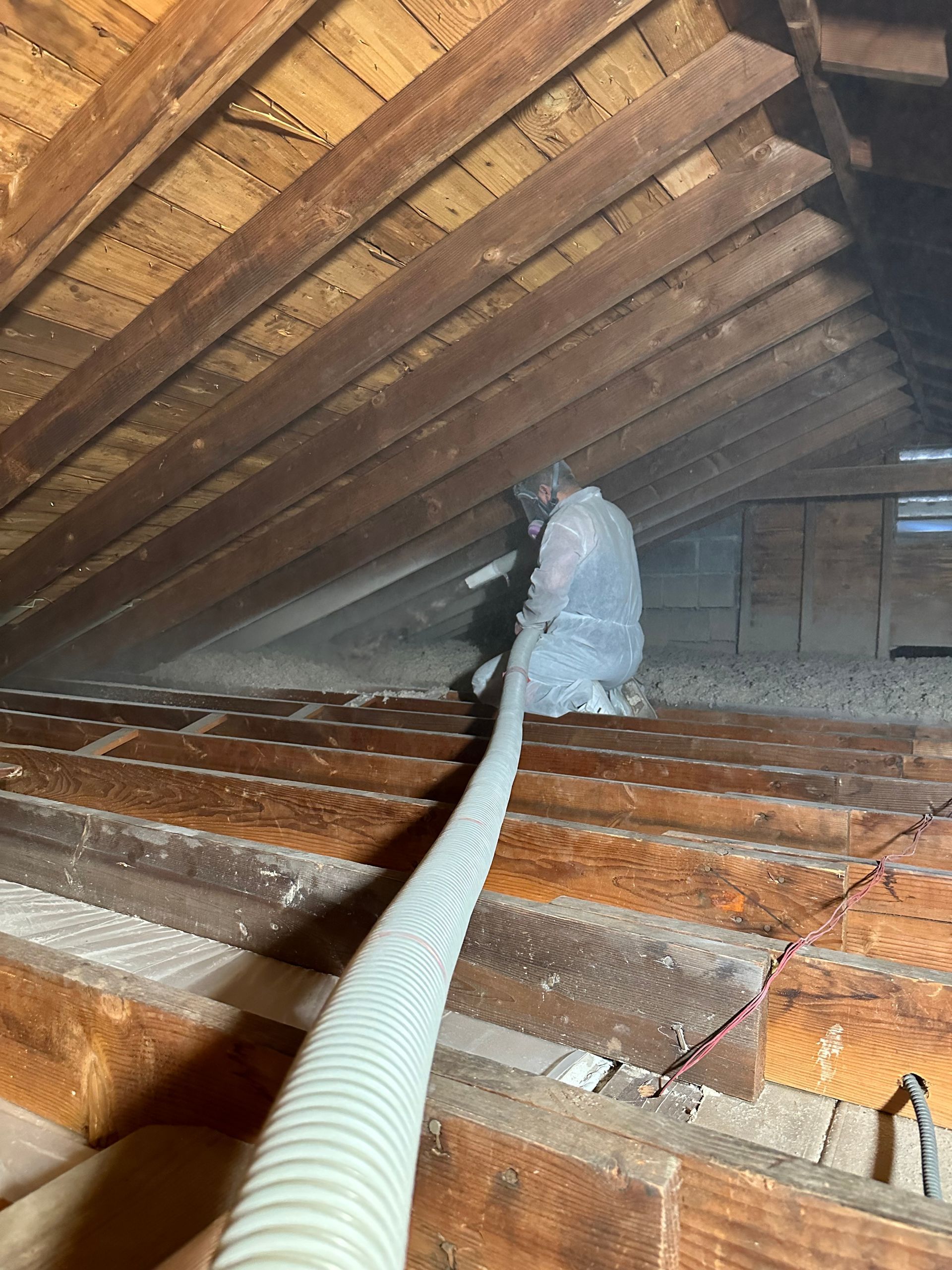 Attic with insulation being installed; a person wearing protective gear handles a hose. Wooden beams are visible.