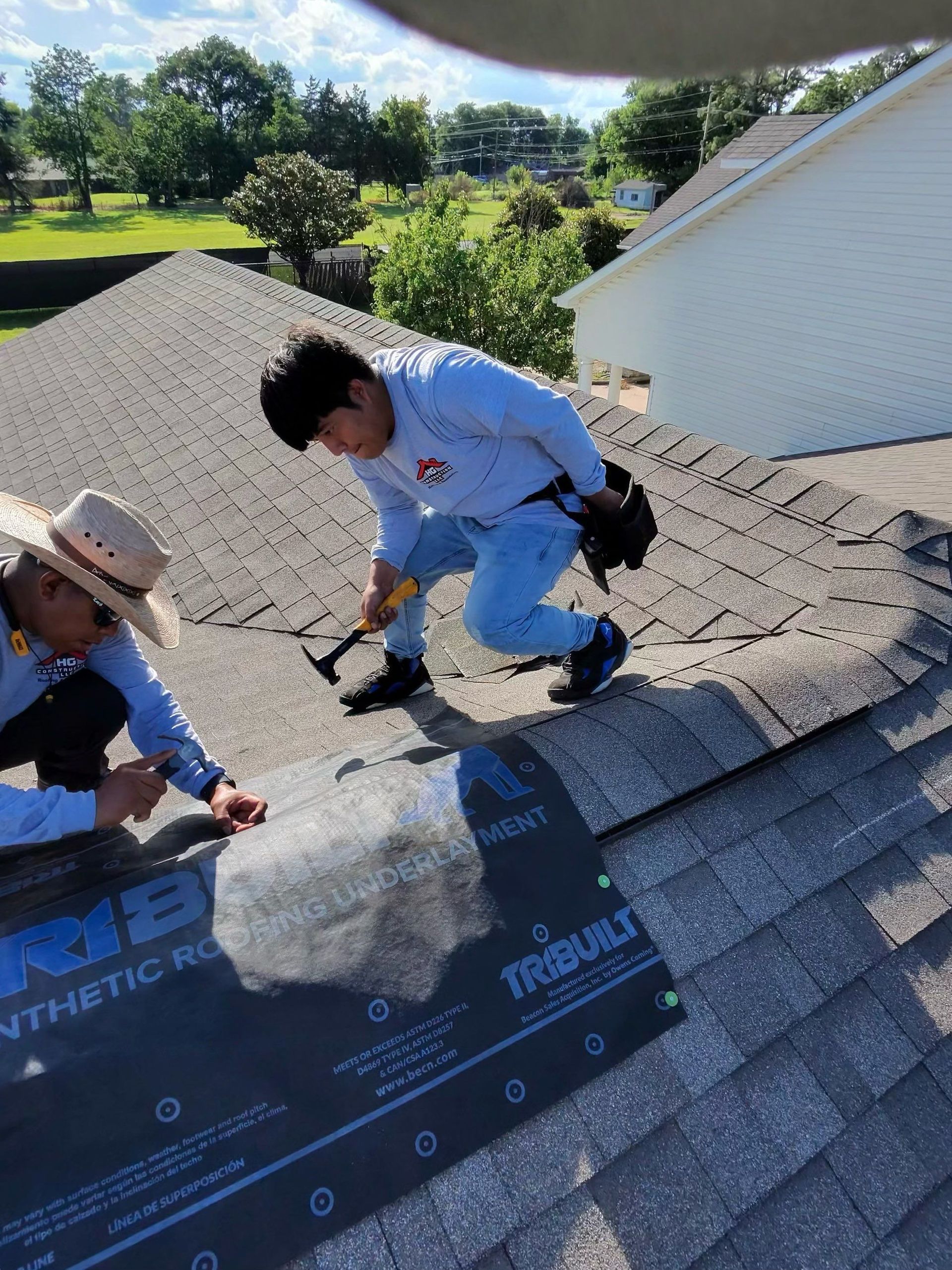 Two roofers on a rooftop, one hammering shingles, another near a synthetic underlayment.