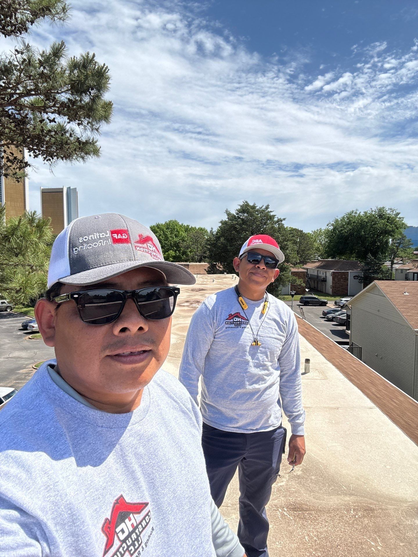 Two men in gray shirts and caps on a rooftop. One takes a selfie, the other stands beside him. Cloudy sky.