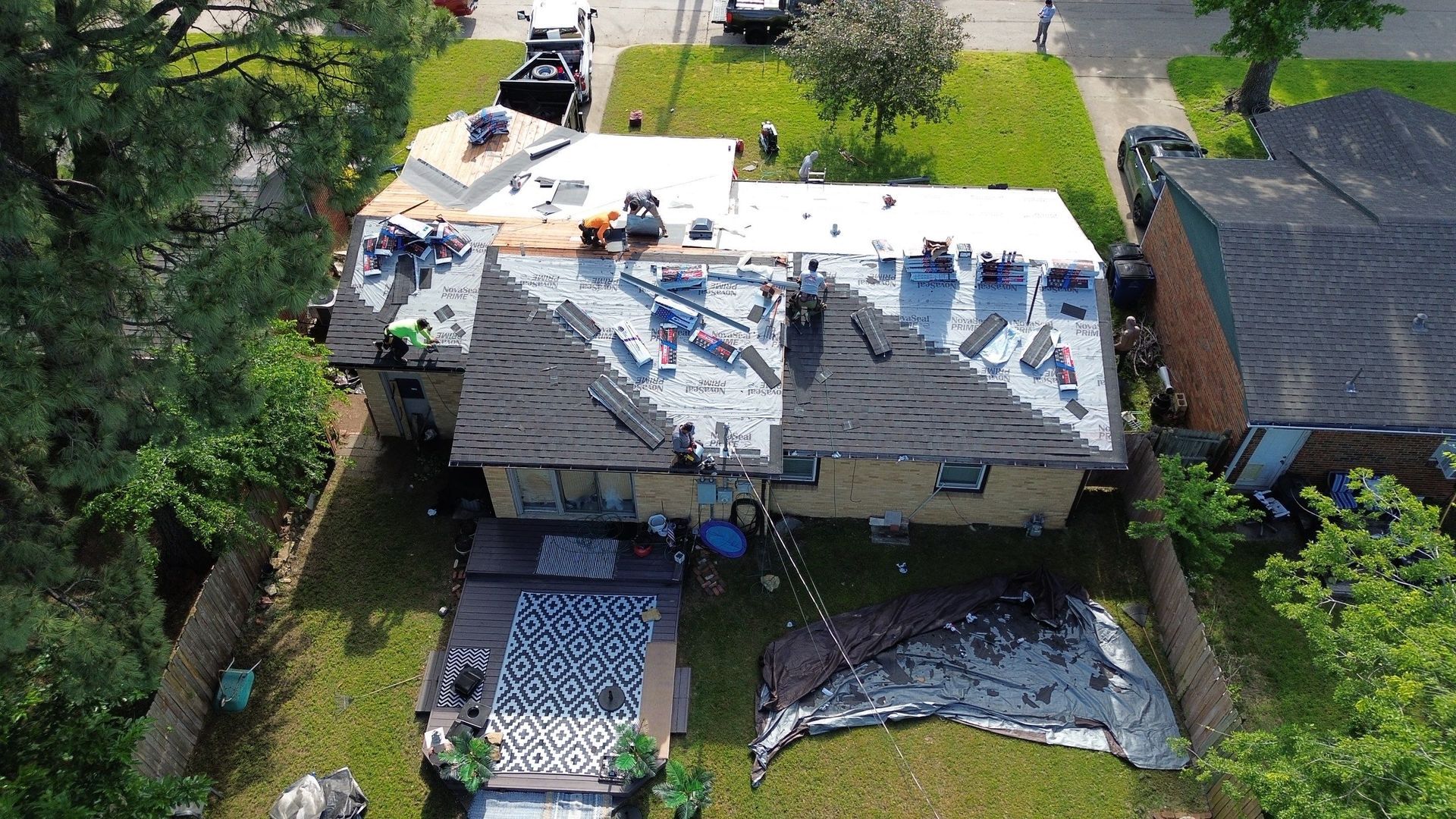 Aerial view of a house roof partially covered with new shingles, workers on the roof, surrounded by trees and grass.