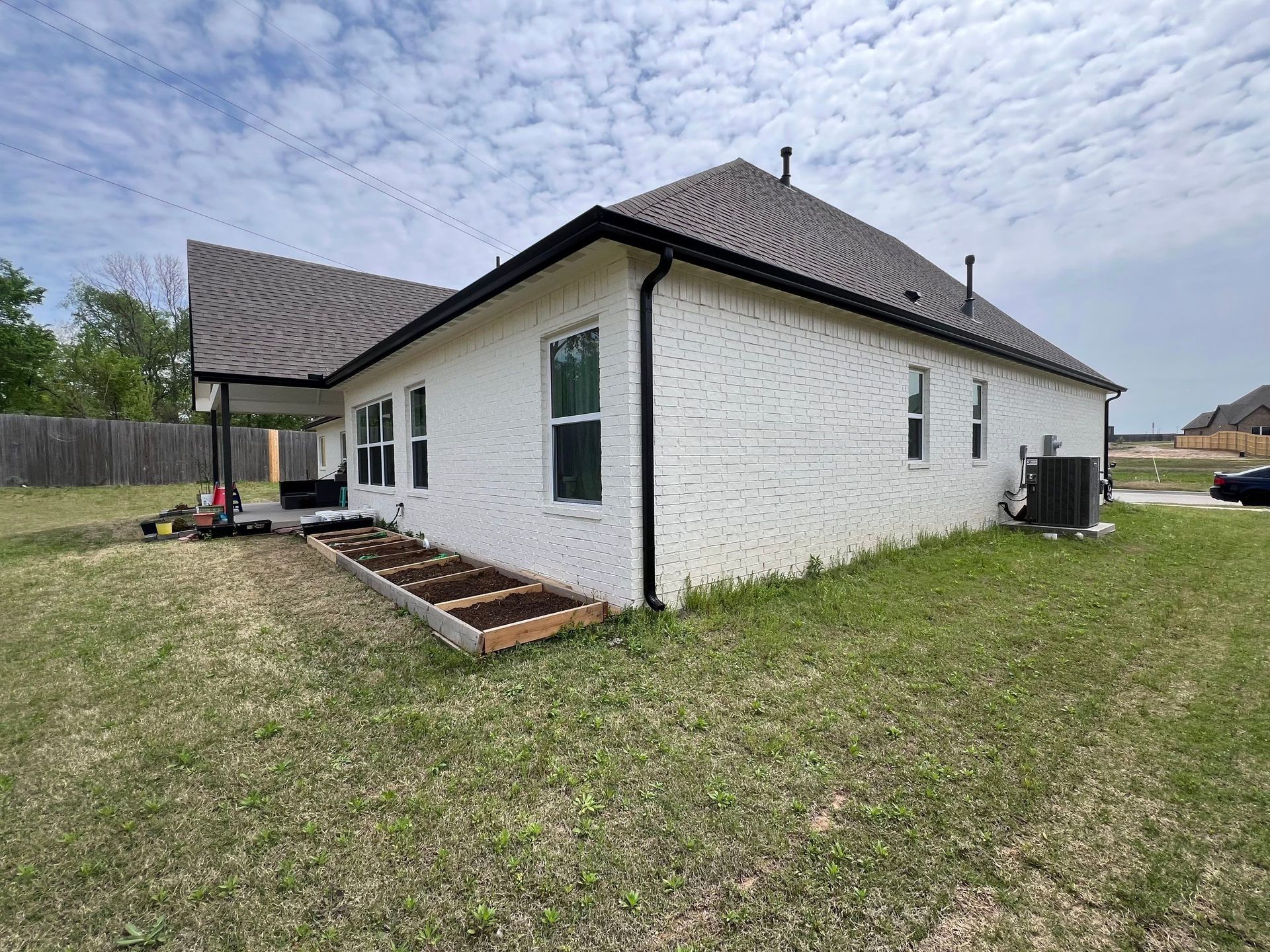 White brick house with black trim, brown roof, and small raised garden bed.