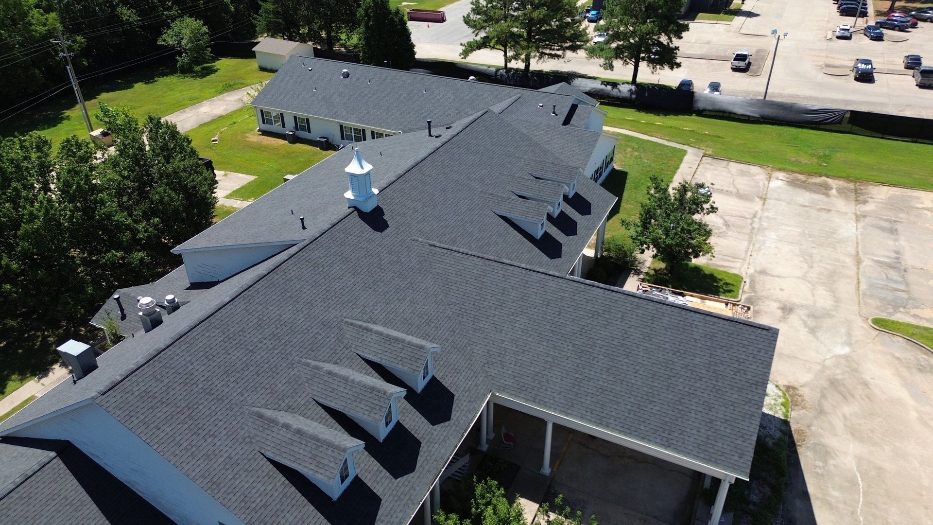Aerial view of a dark shingled roof with a white cupola and dormers, trees, and a parking lot.