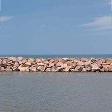 A horizontal line of jagged, light brown rocks separates calm, grey water from a clear blue sky.