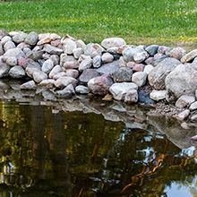 A pile of rounded grey and tan stones lining the edge of a calm, reflective pond with grass in the background.