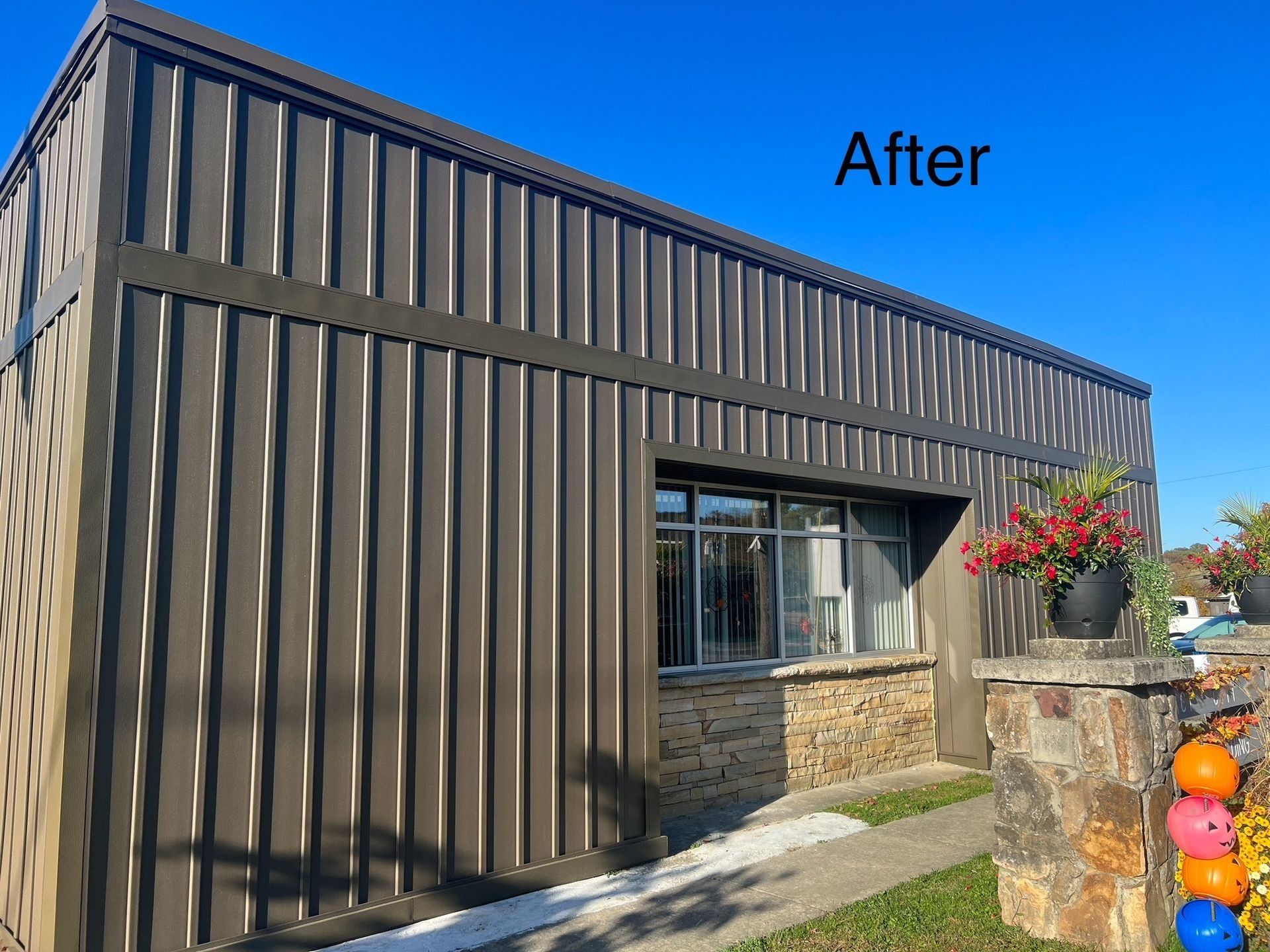 Brown metal-sided building with window, stone base, flower pots, and blue sky.