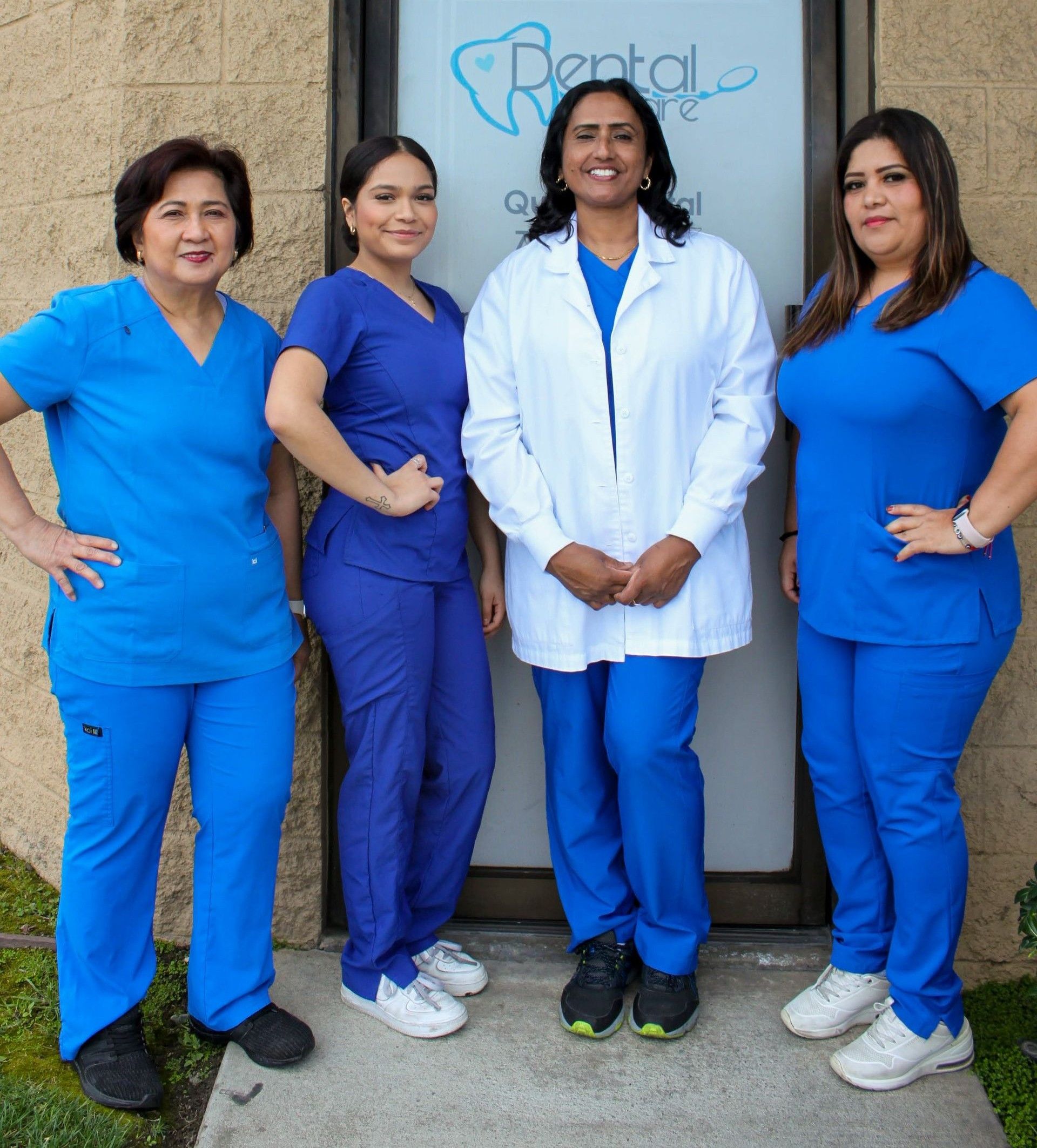 Four dental staff in blue scrubs and a white coat pose in front of a clinic entrance.