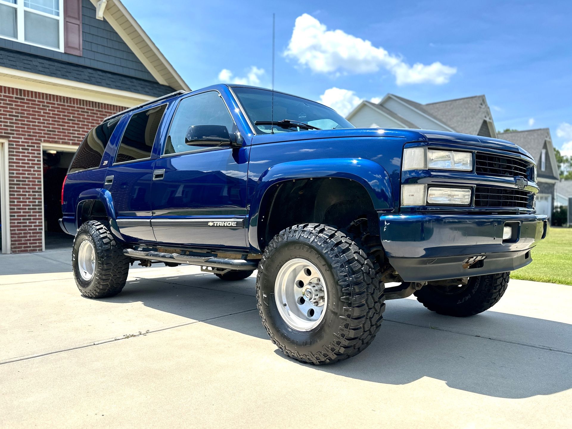 Blue lifted Chevy Suburban SUV parked outside a brick home on a sunny day.