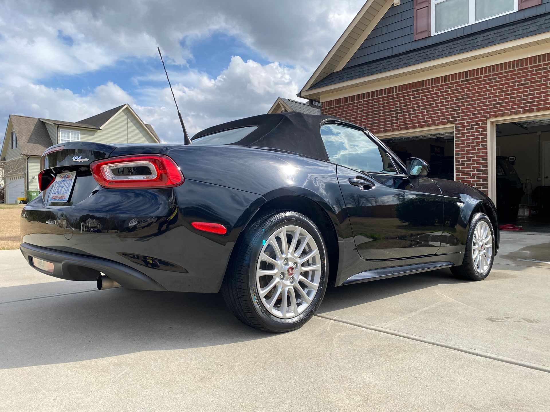 Black convertible car parked in front of a brick house with a partially open garage on a sunny day.