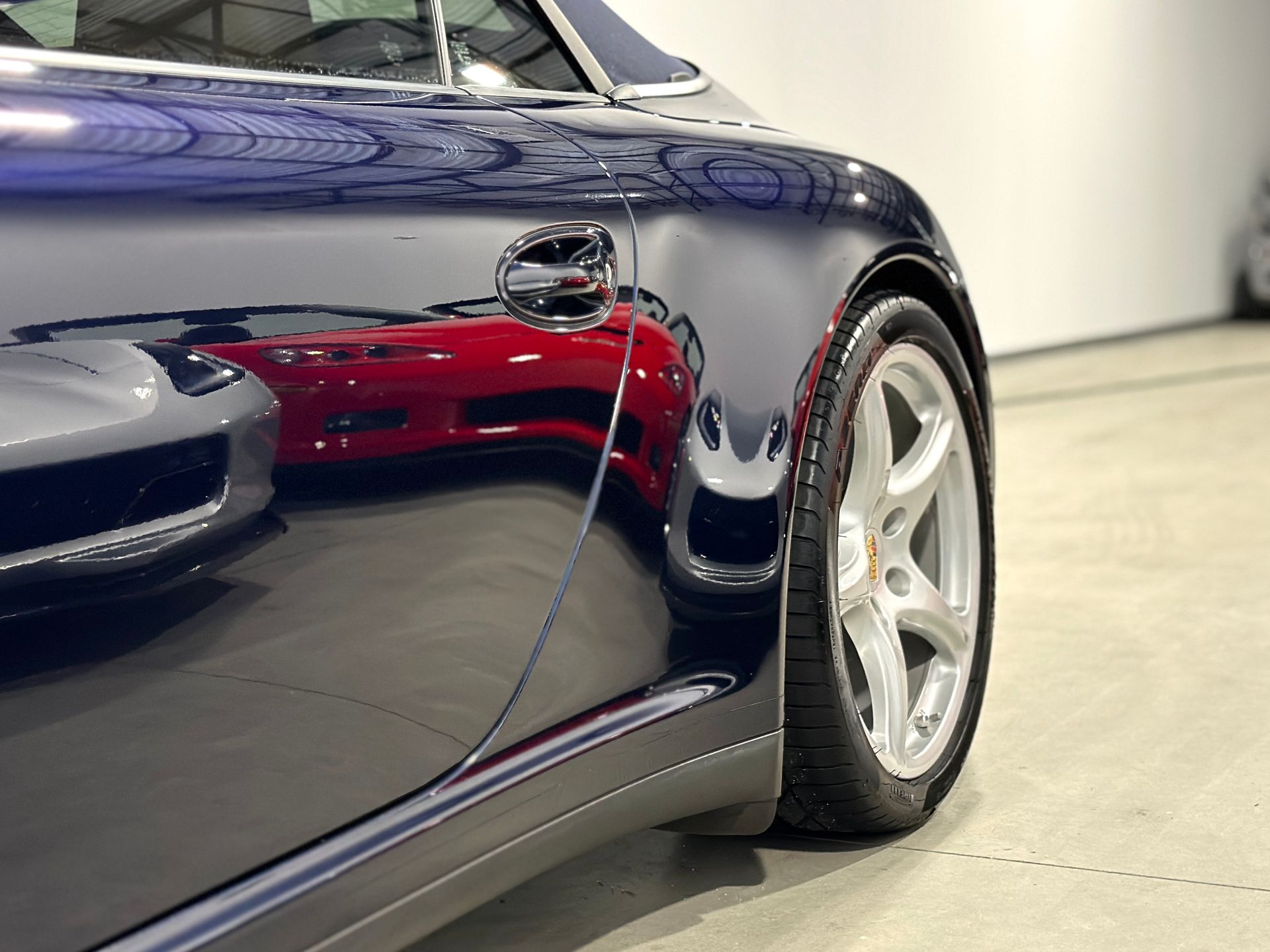 Dark blue sports car's side, reflecting a red car, silver wheel, in a well-lit garage.