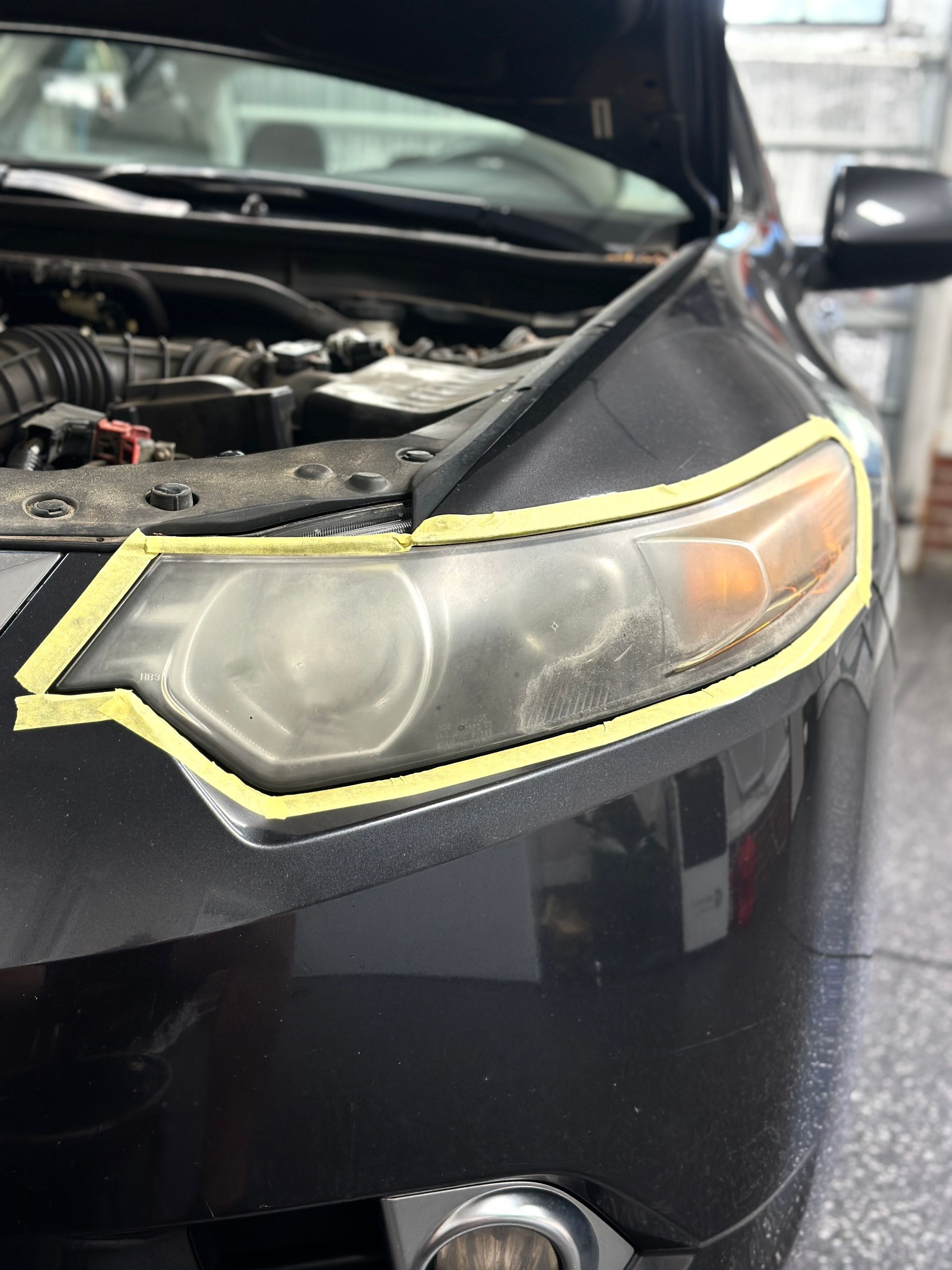 Black car's headlight with yellow tape around it, prepping for restoration. Hood open, setting is a garage.