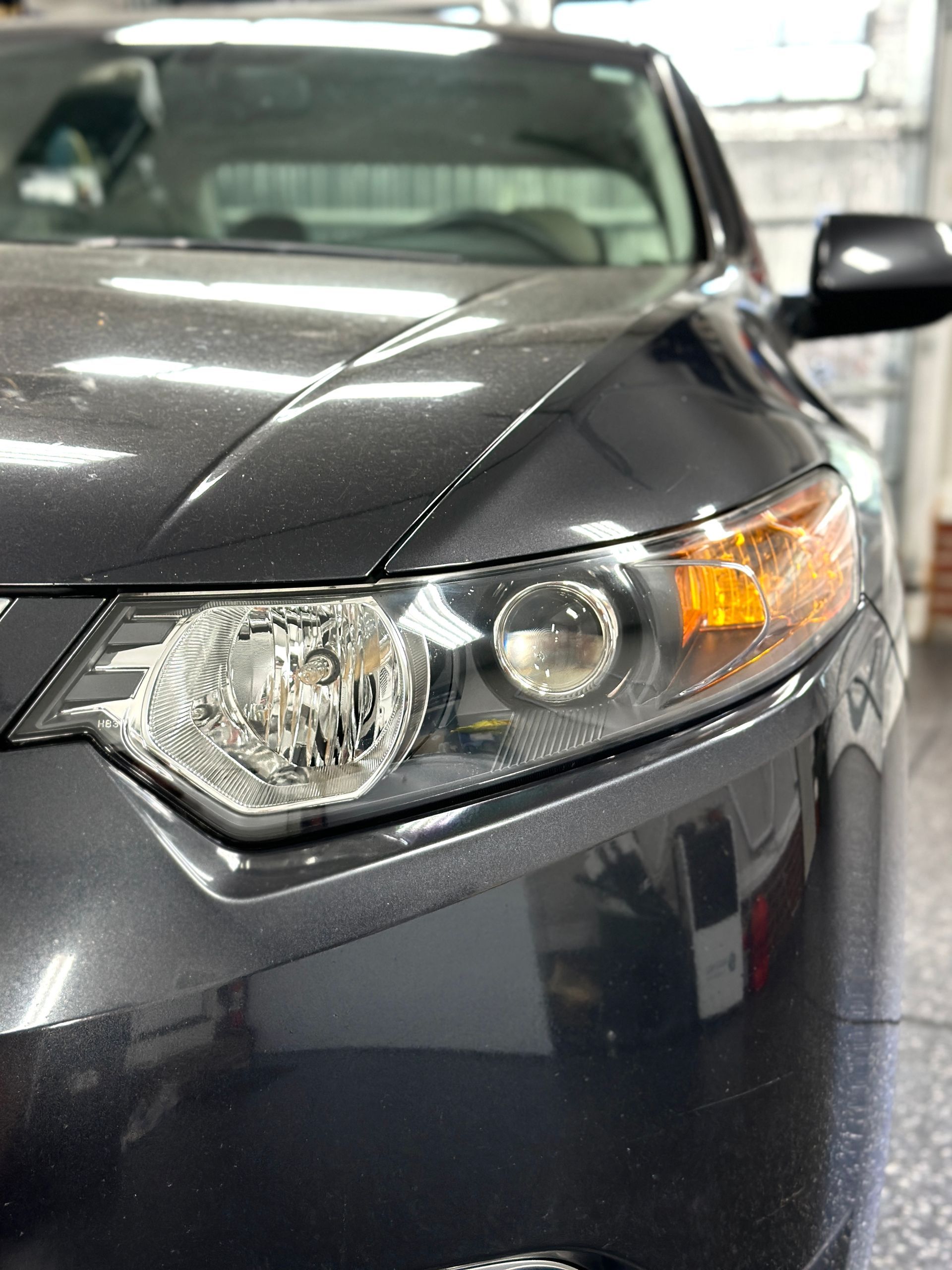 Close-up of a dark gray car's front with a headlight and side marker light visible, parked indoors.