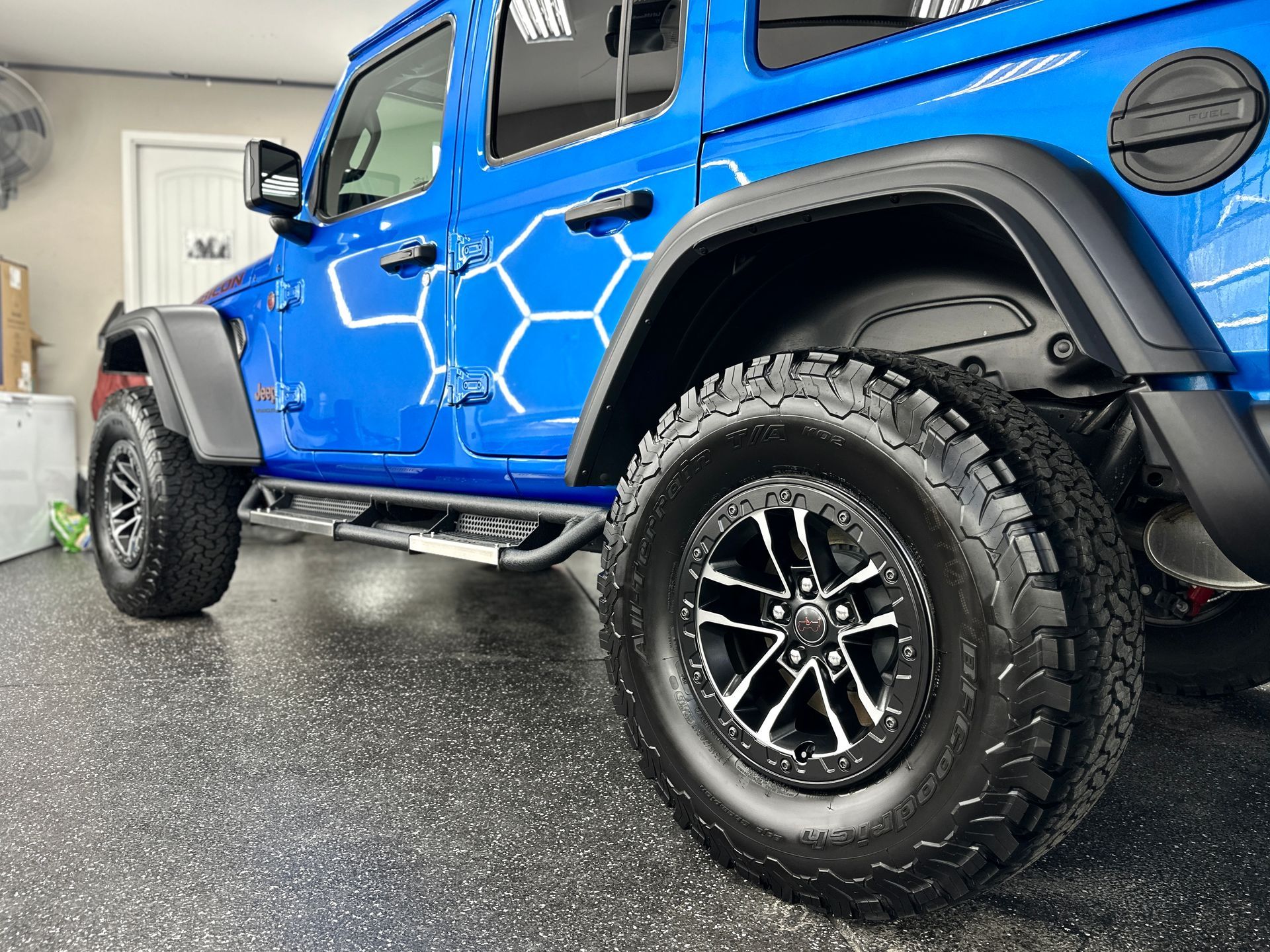 Blue Jeep with black wheels and tires parked inside a garage; hexagonal design on the side.