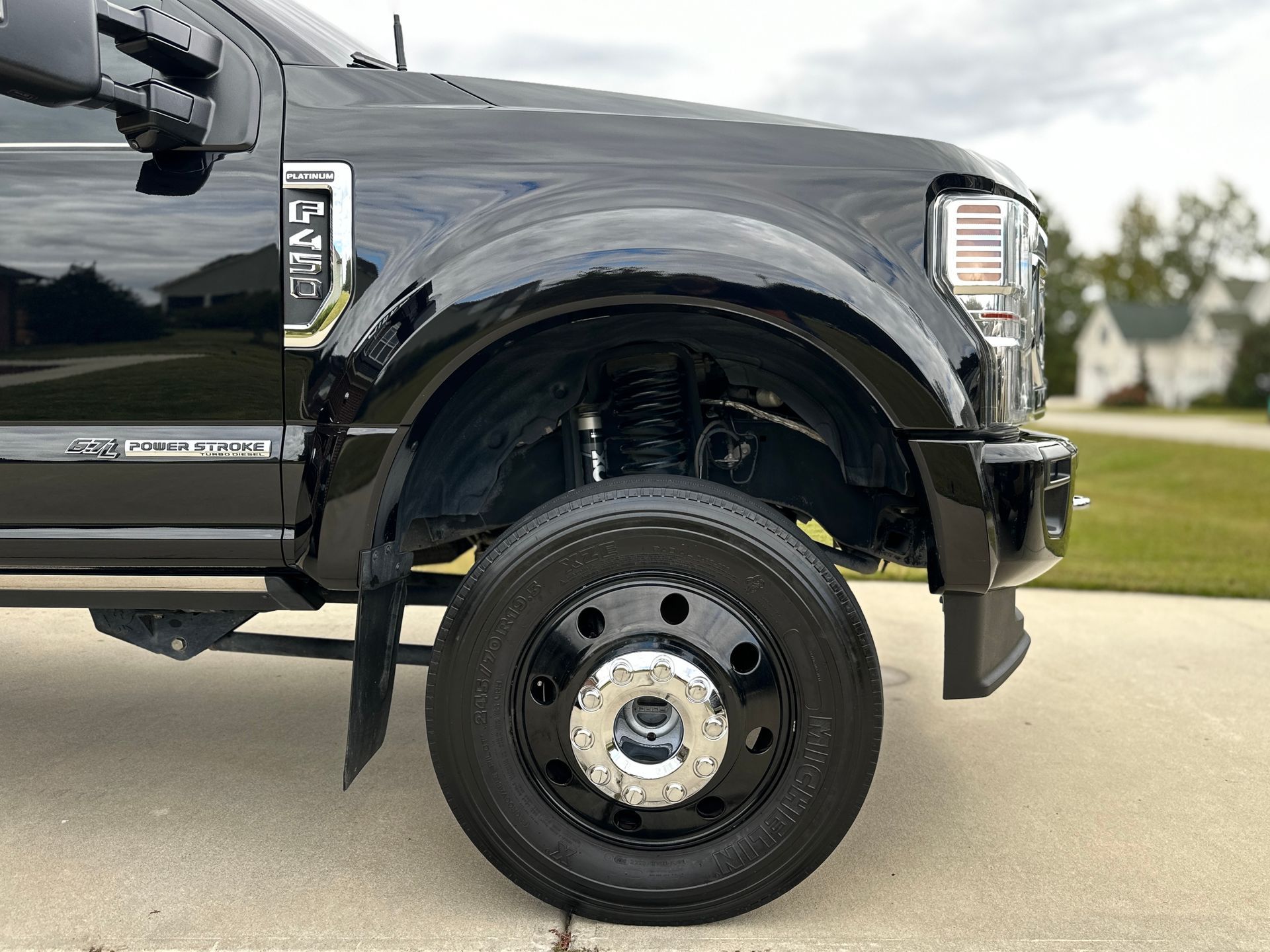 Black Ford truck's front wheel, parked on a driveway. Shiny black paint, chrome hubcap, and visible suspension.