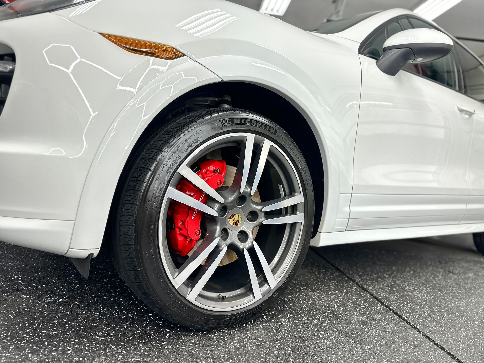 White Porsche SUV with a red brake caliper, tire, and wheel in a garage.