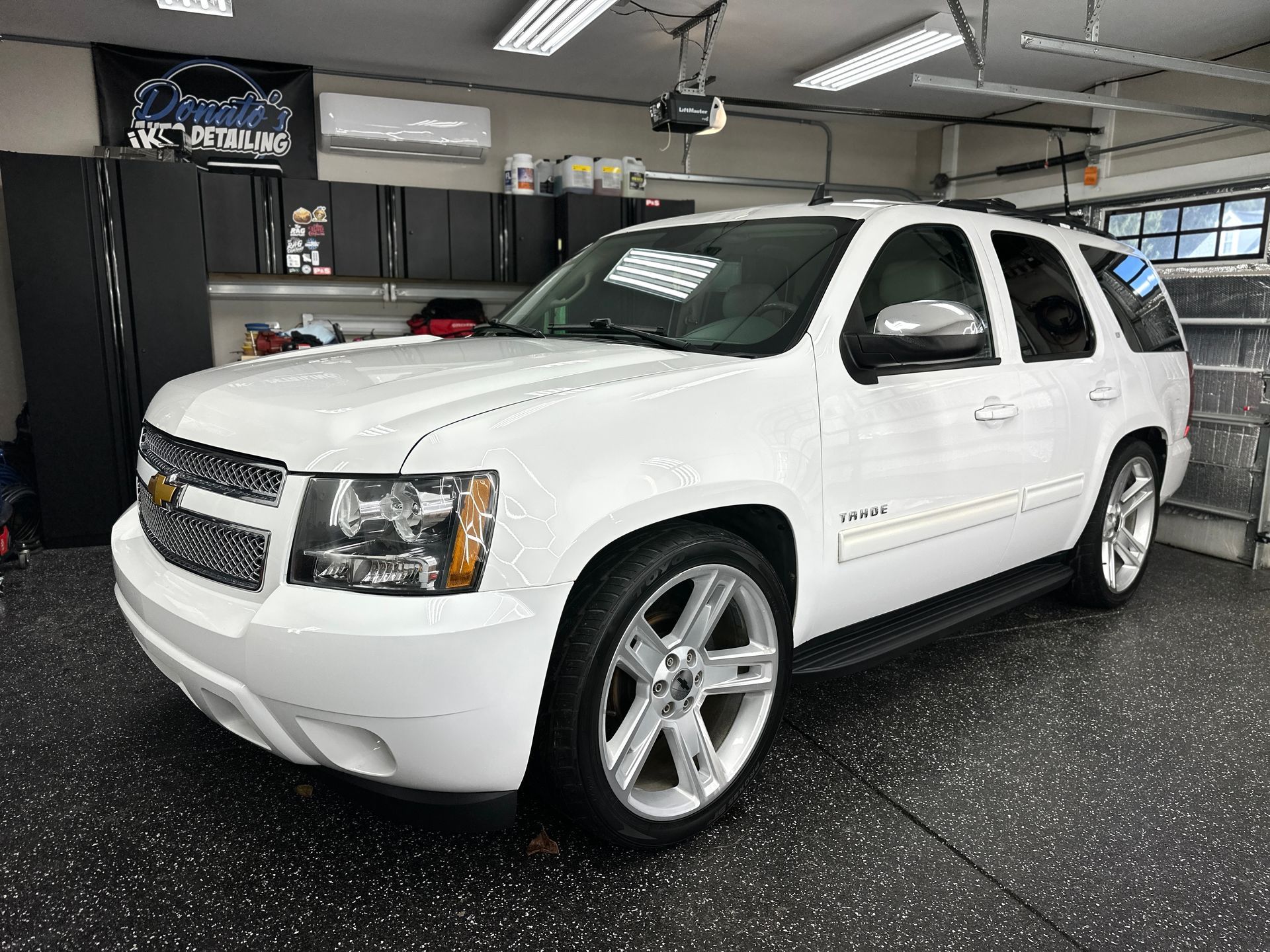 White Chevrolet Tahoe SUV parked inside a garage with black cabinets and speckled flooring.