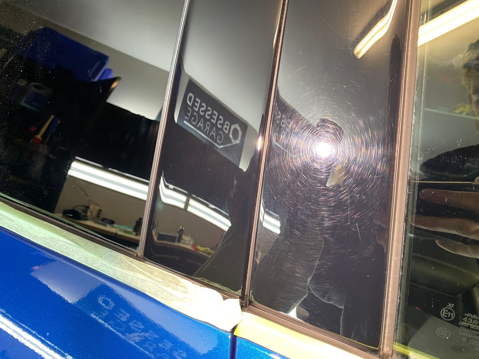 Close-up of a blue car's window with visible scratches and reflections. Workshop setting.