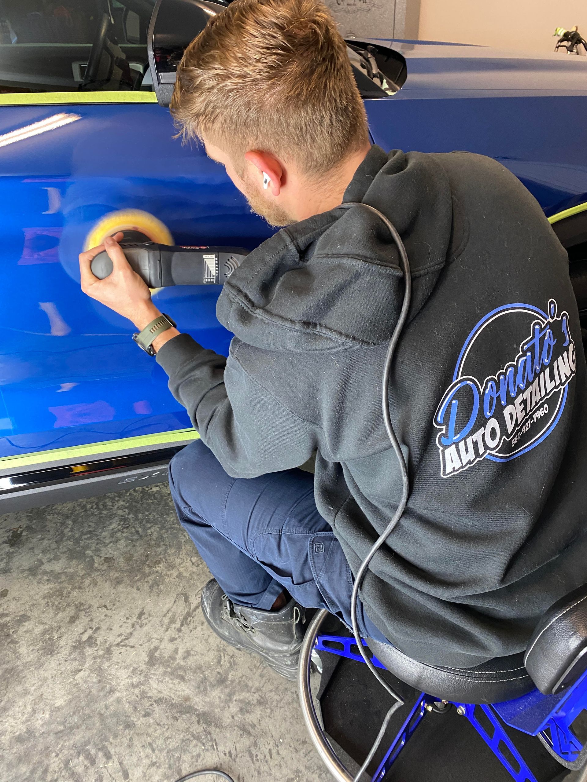 Man polishing blue car with power buffer in a garage.