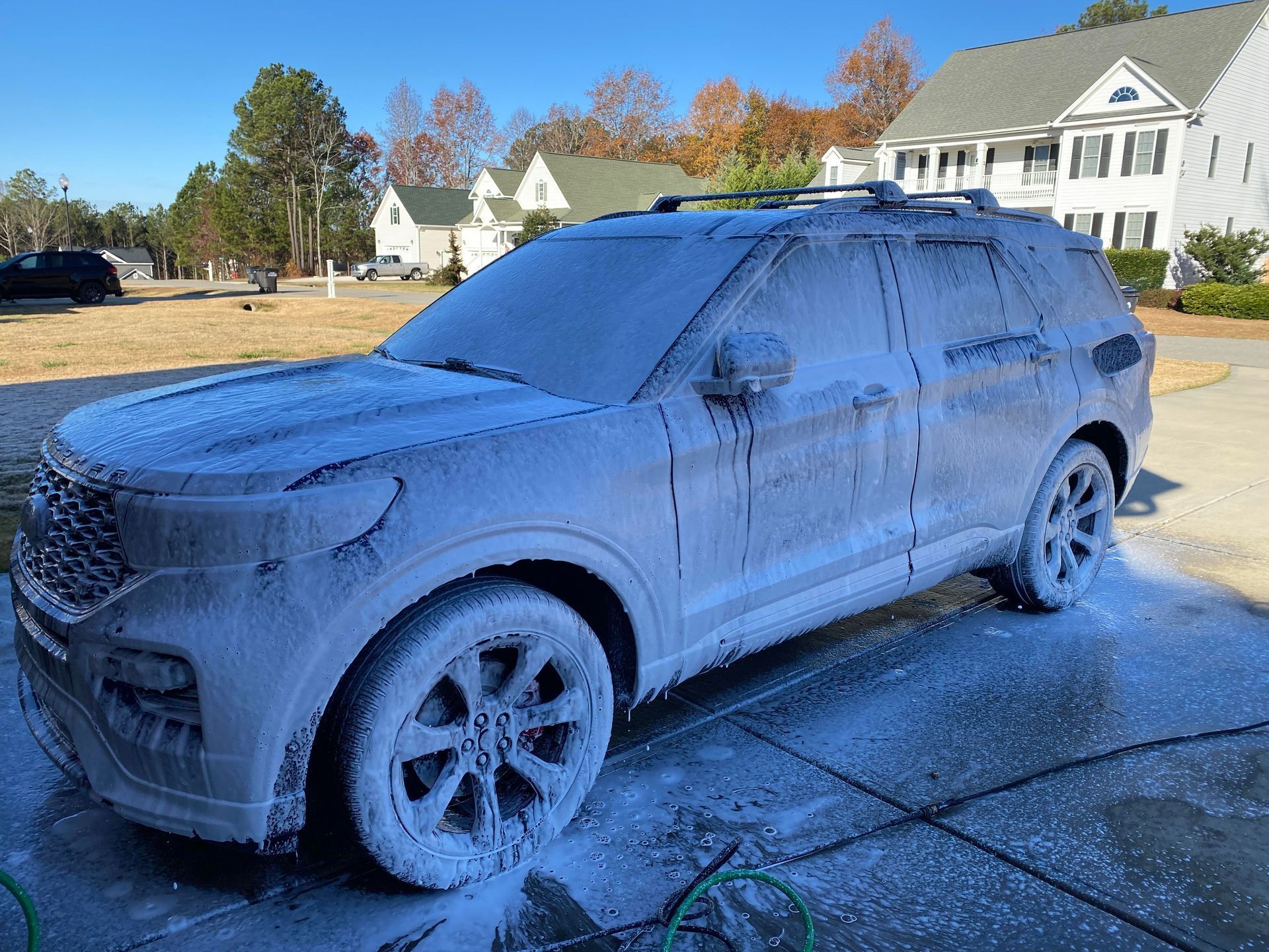 SUV covered in white foam suds, being washed in a driveway on a sunny day.
