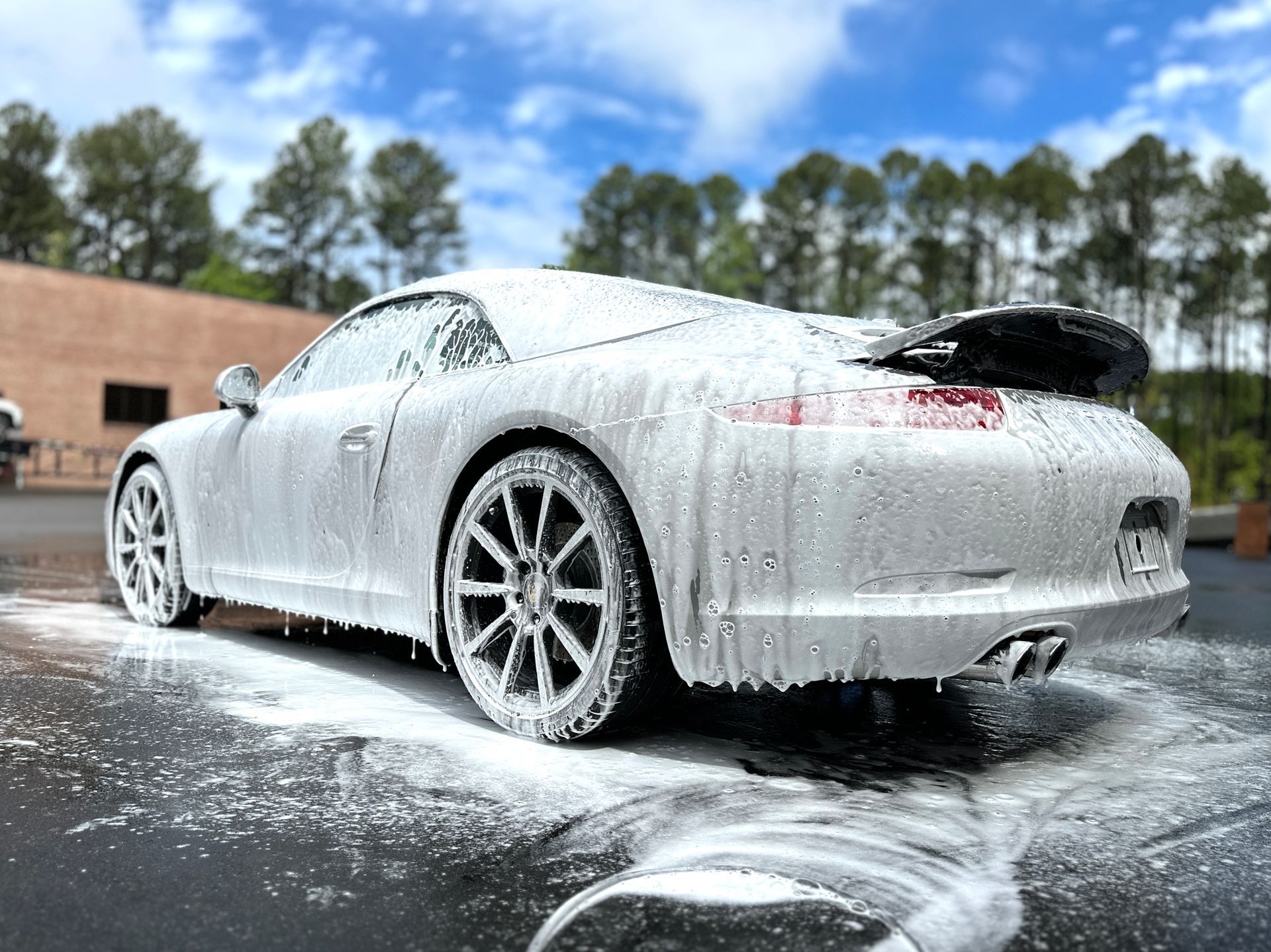 White sports car covered in car wash foam outdoors on a sunny day.