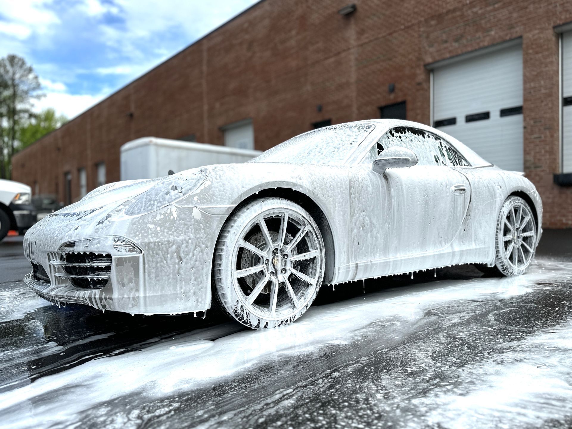 A silver sports car covered in white foam suds, parked outside a brick building.