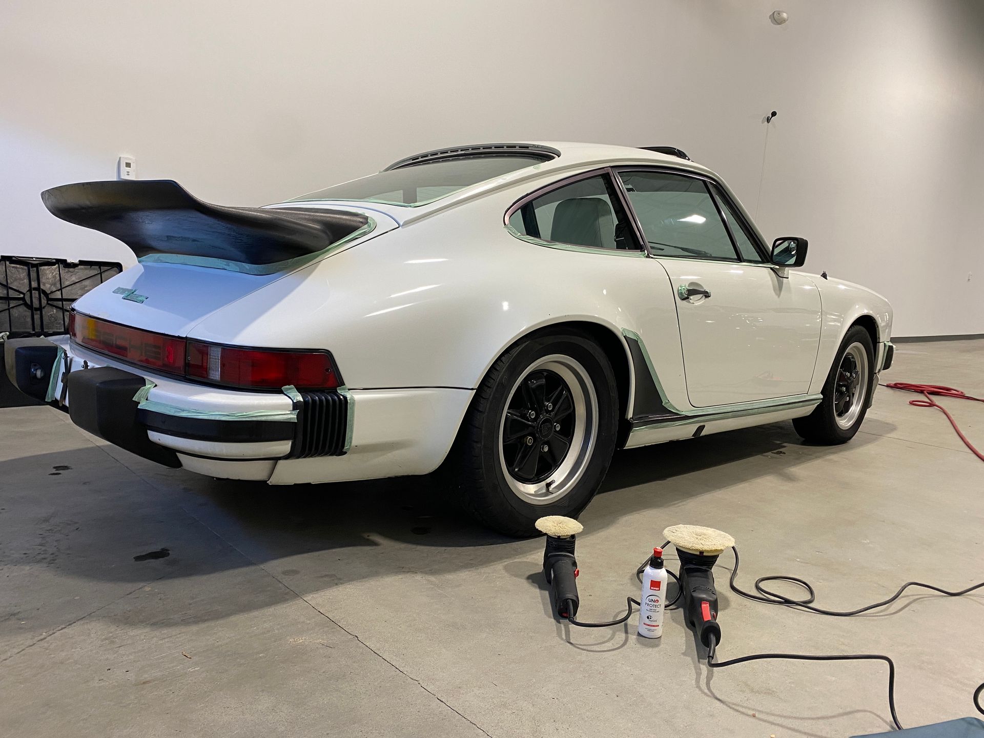 White Porsche 911 with black wheels and spoiler, in a garage with polishing tools.