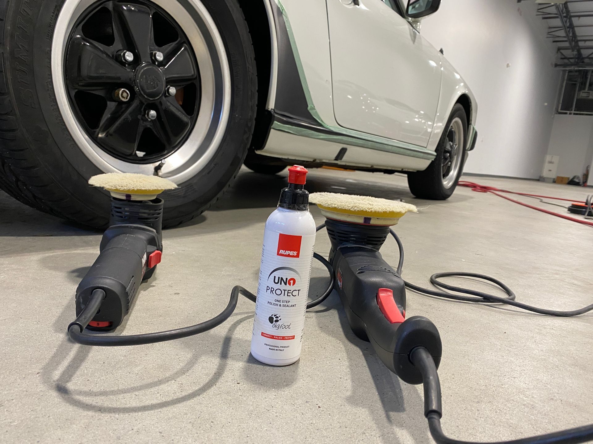 Two car polishers and a bottle of polish by a white car with black wheels.
