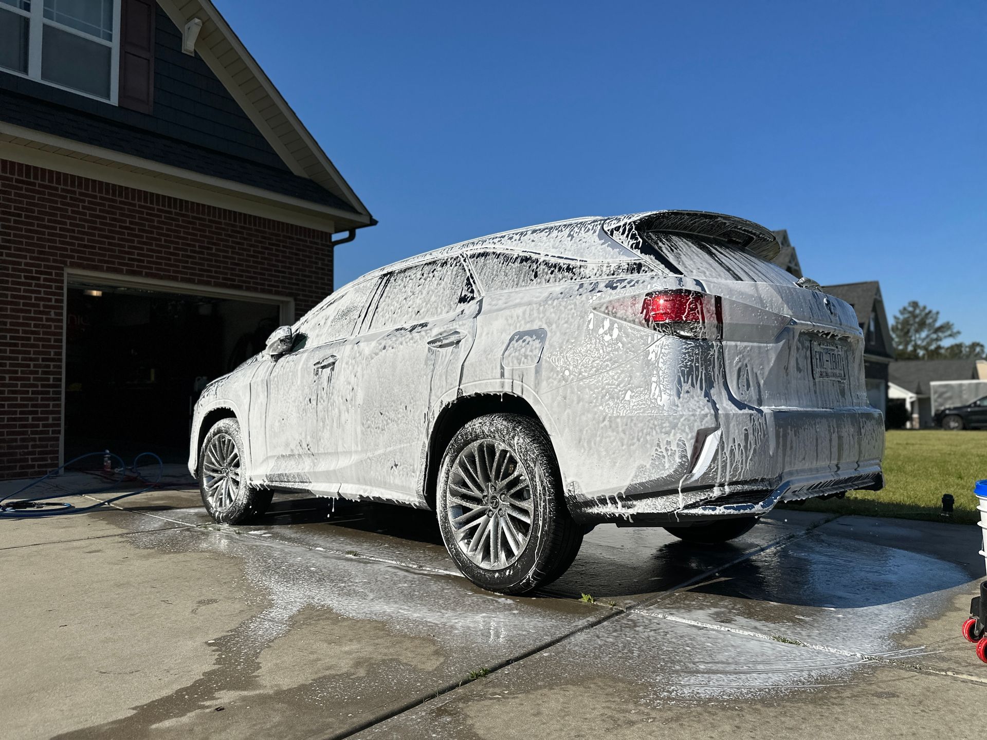 A white SUV covered in foamy soap suds sits on a driveway in front of a house, car wash in progress.