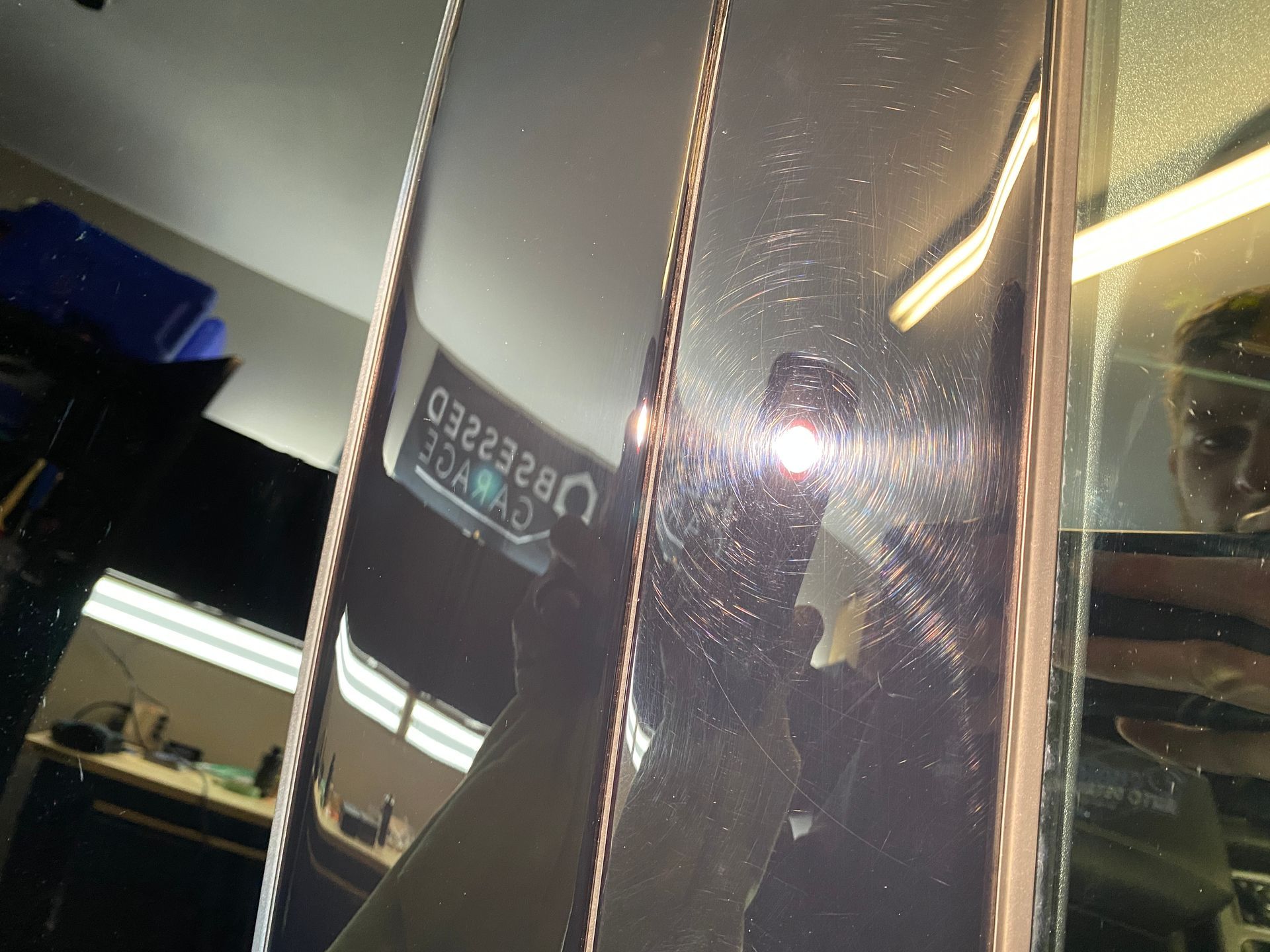 Close-up of a dark car panel with visible scratches reflecting light. A person holding a phone is reflected in the panel.
