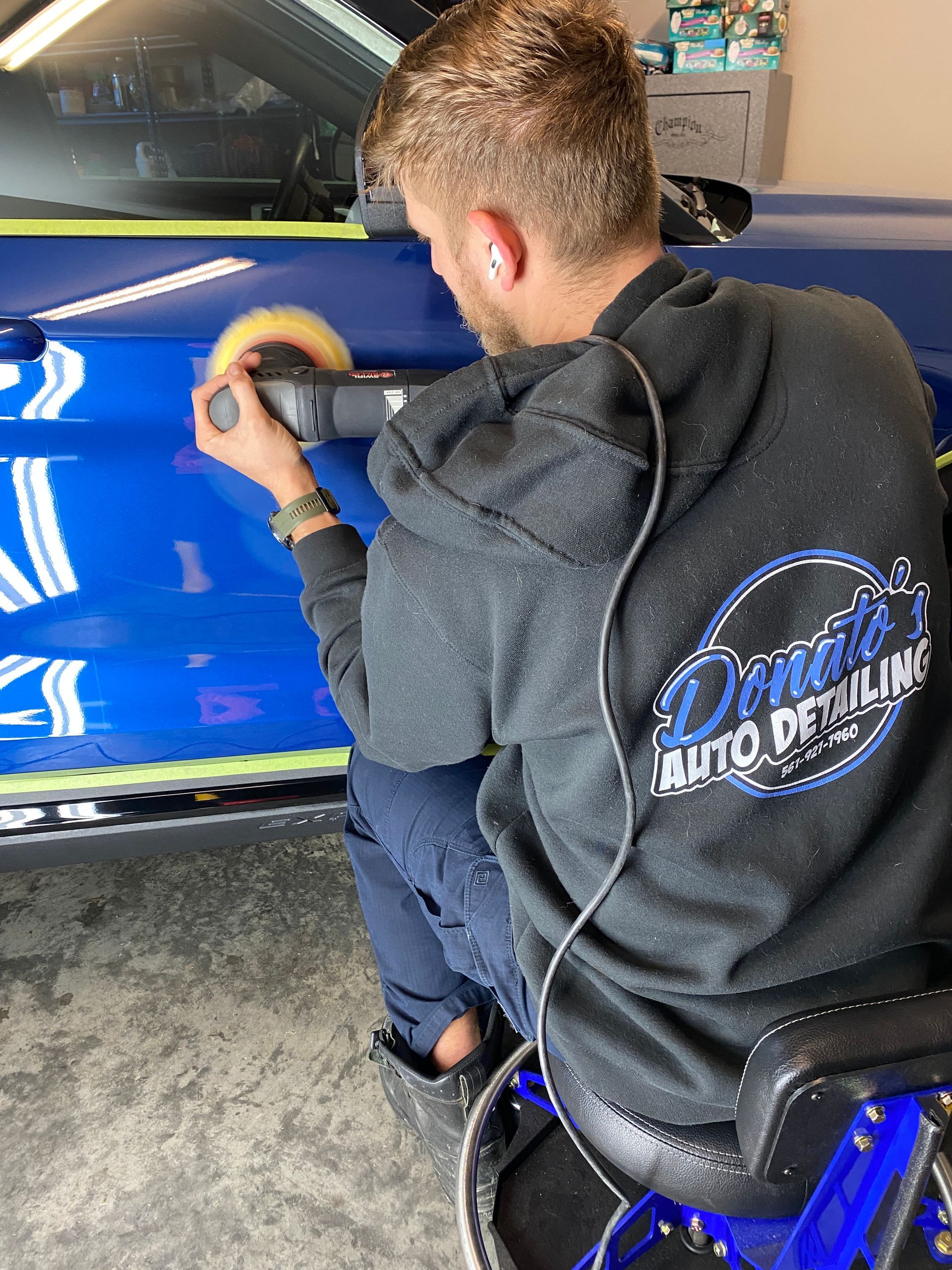 Man polishing blue car door with power tool. He wears a black hoodie with logo 
