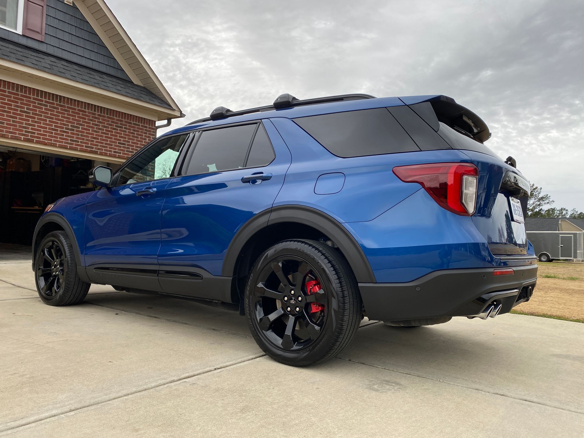Blue Ford Explorer SUV with black wheels and roof rack parked in front of a house on a cloudy day.