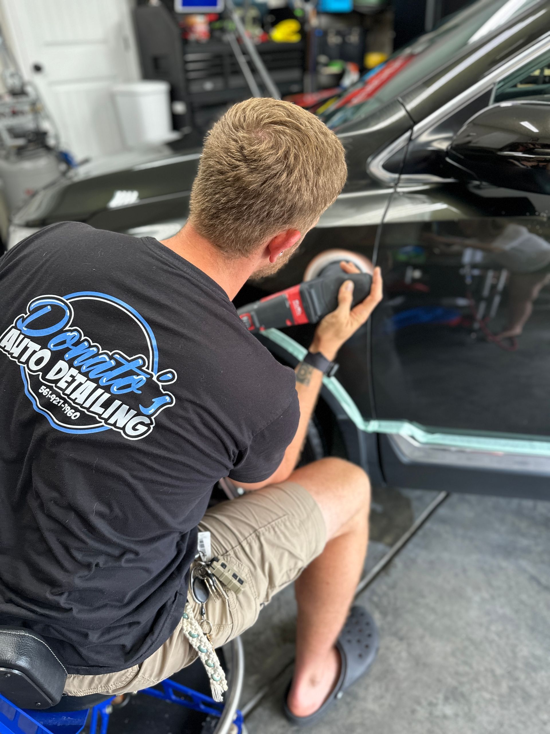 Man polishing a black car door with an electric polisher in a garage.