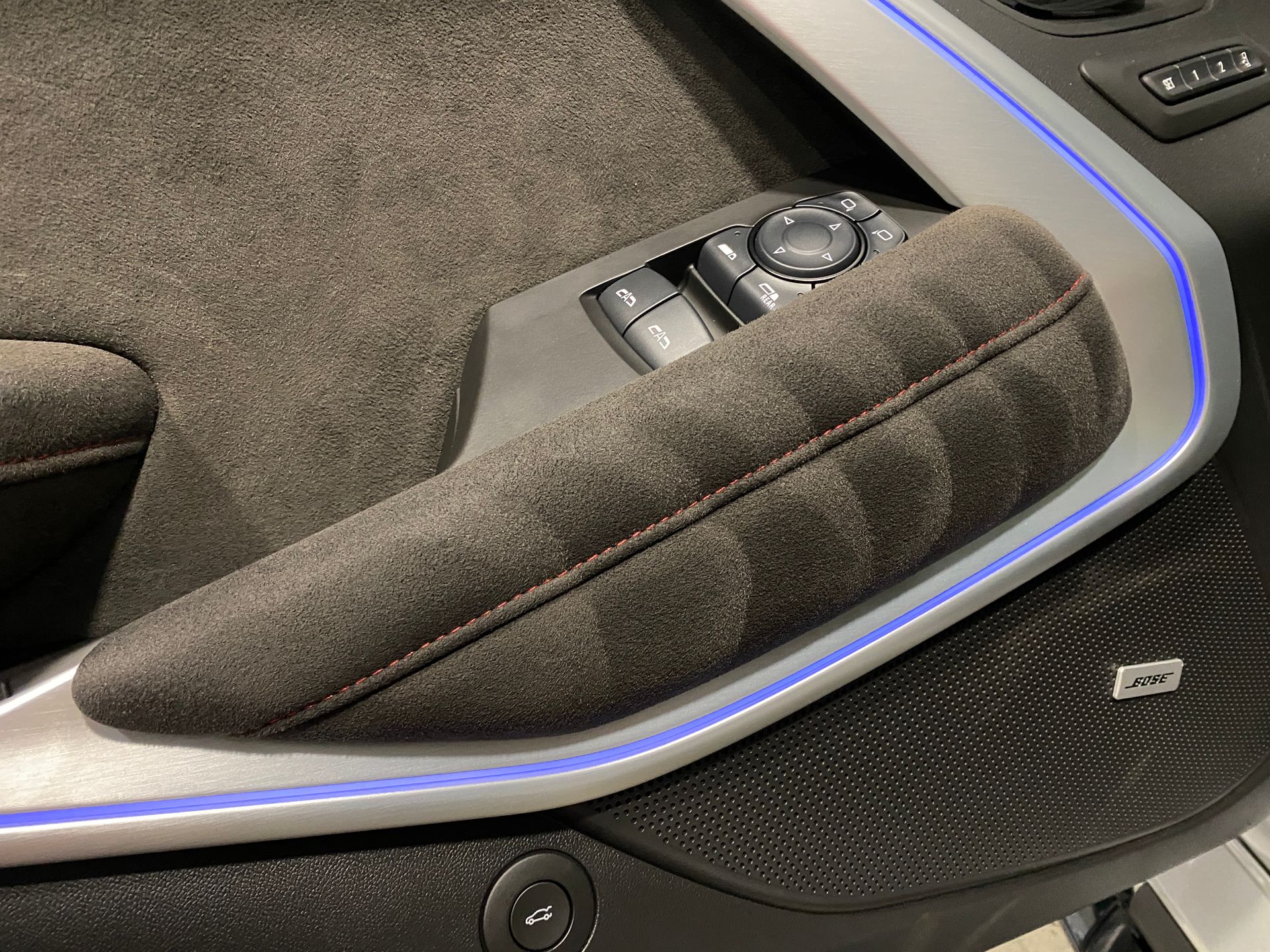 Car door interior, suede armrest with red stitching, ambient blue light, and control panel.
