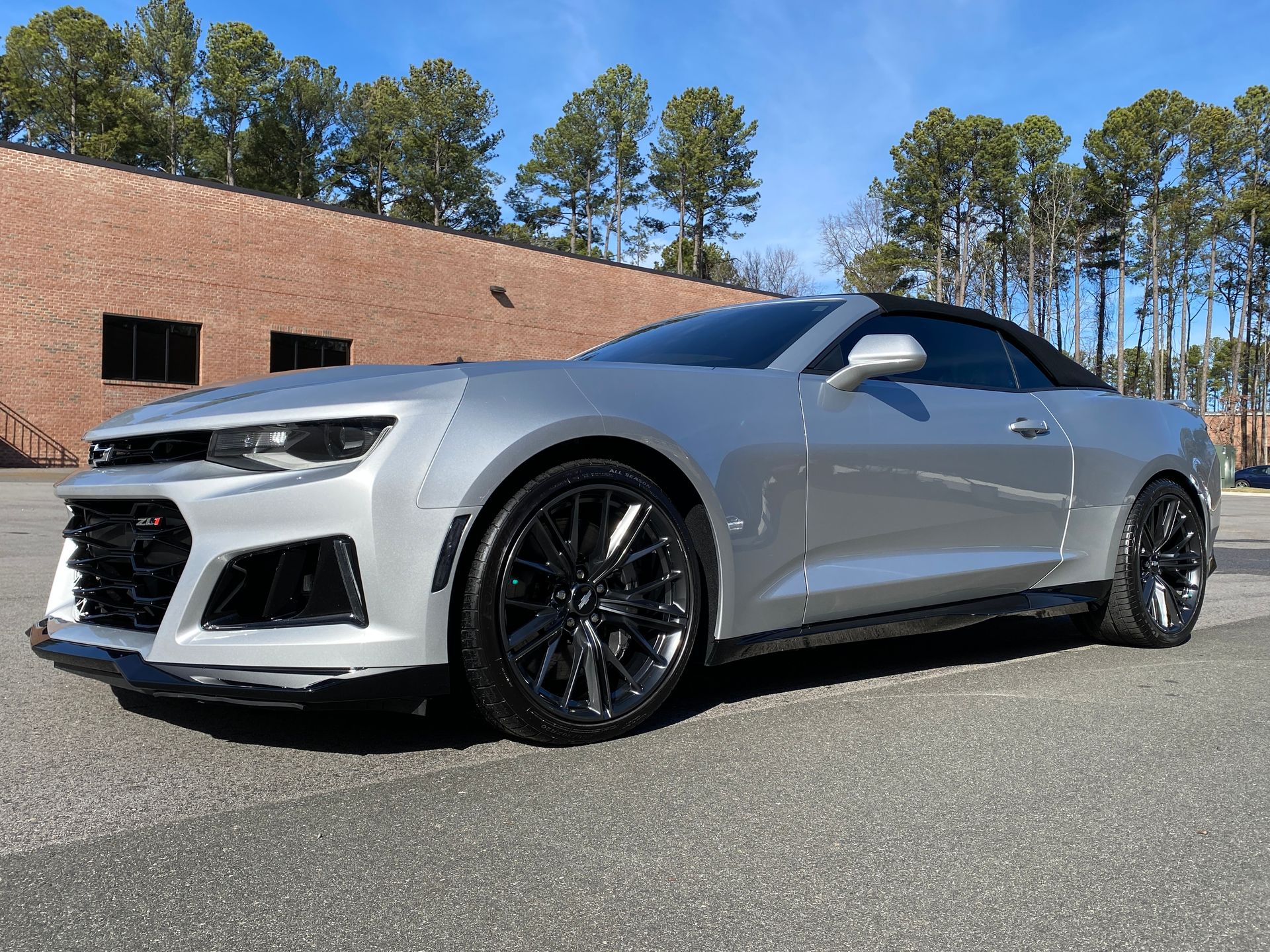 Silver convertible Chevrolet Camaro parked on asphalt. Black wheels, dark top, brick building background.