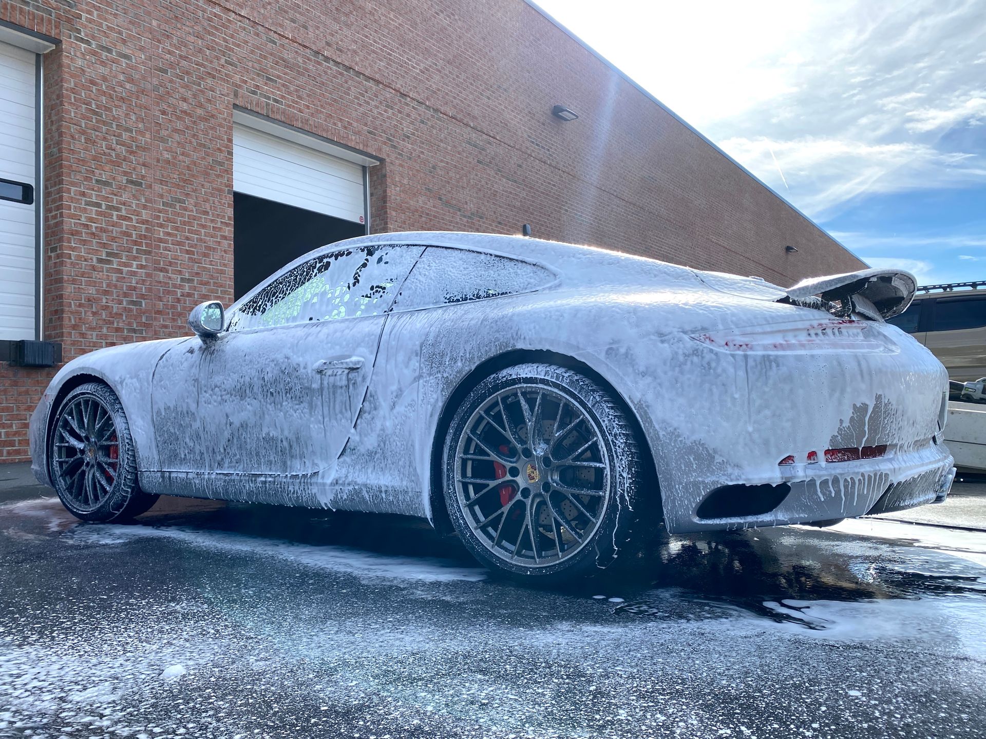 Silver Porsche covered in white foam, being washed outside a brick building.