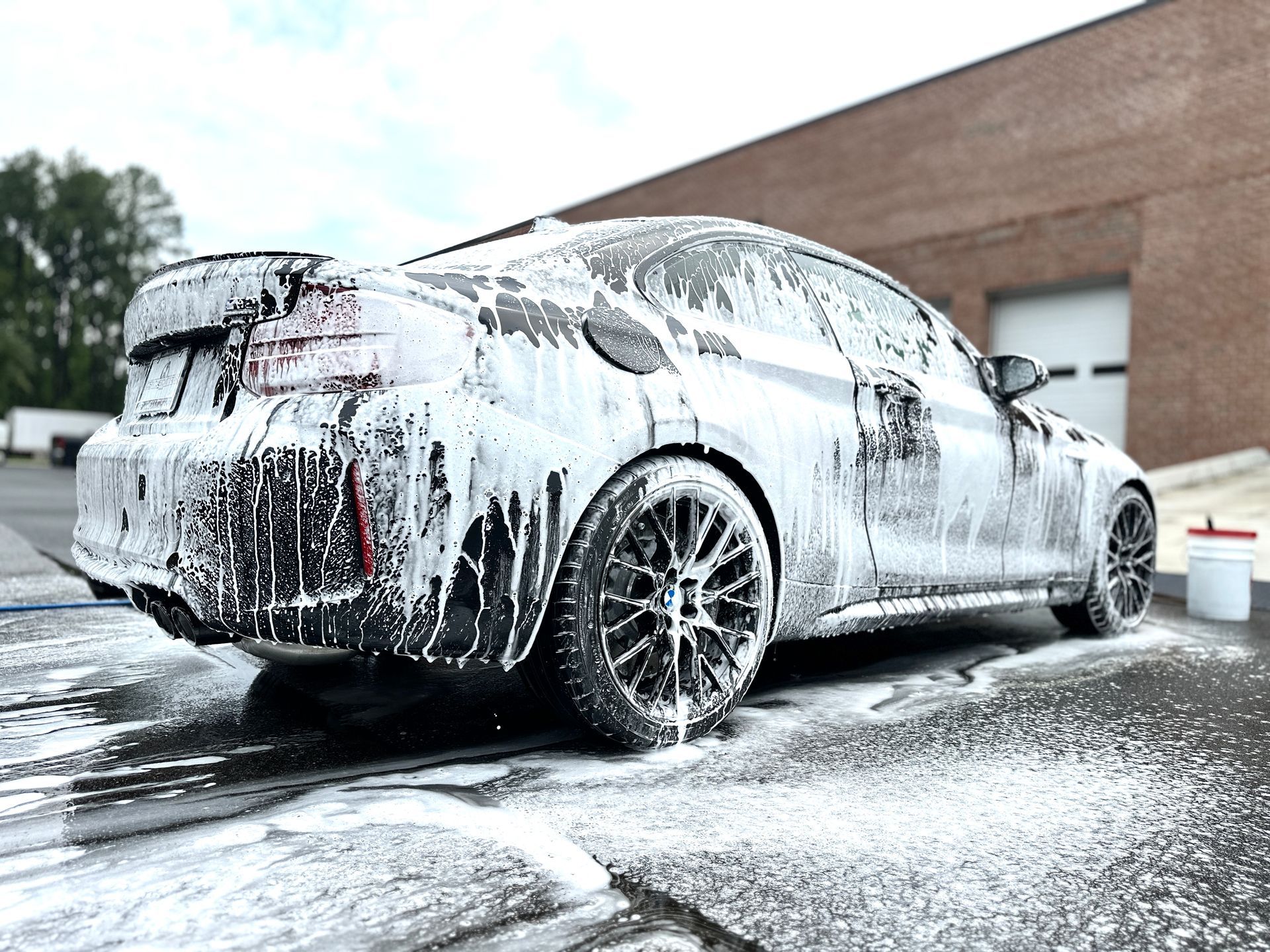 Black car covered in white foam, being washed outside a brick building.