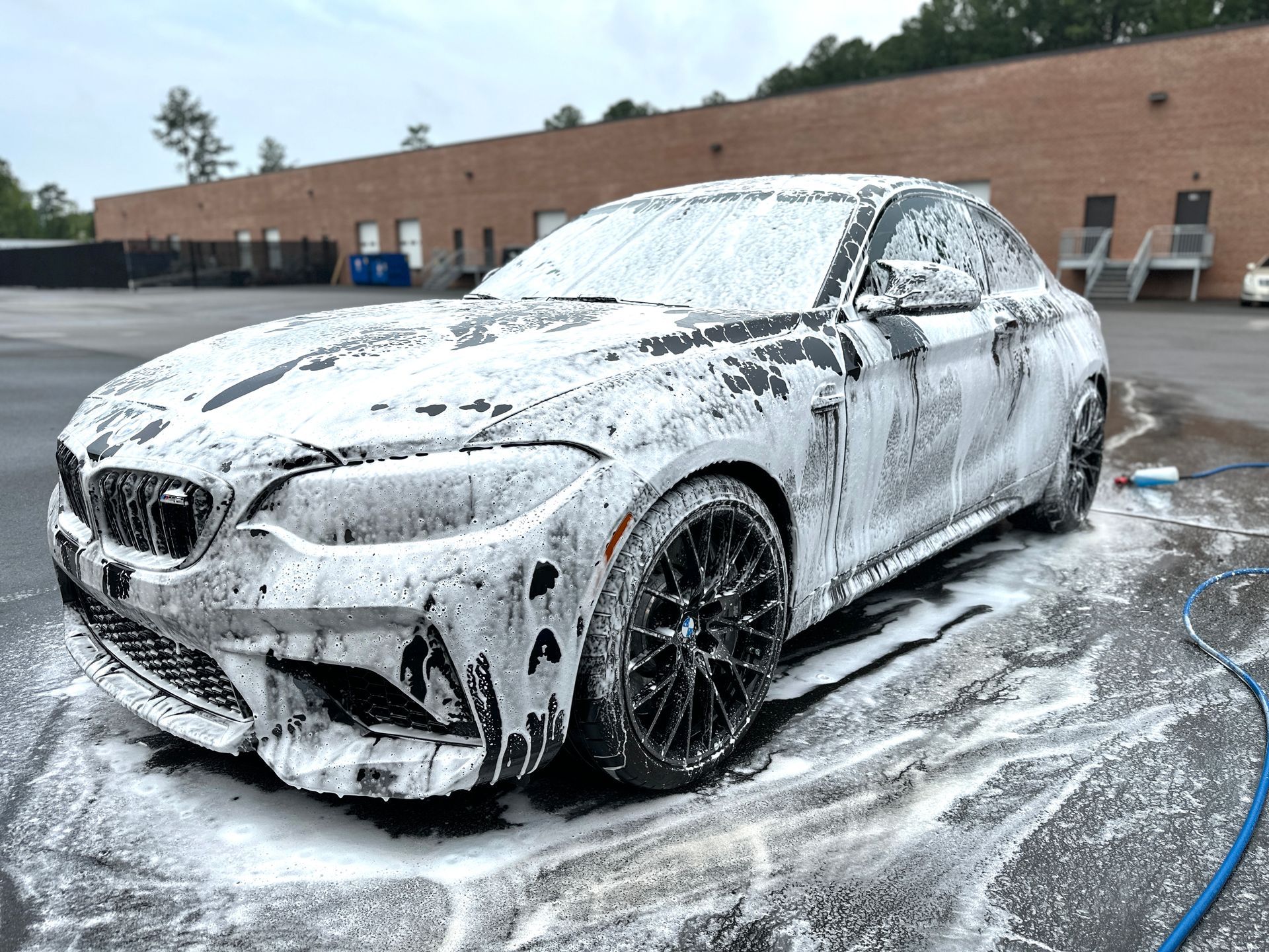 Black BMW coupe covered in white foam, undergoing a car wash. Outdoors, near a building.