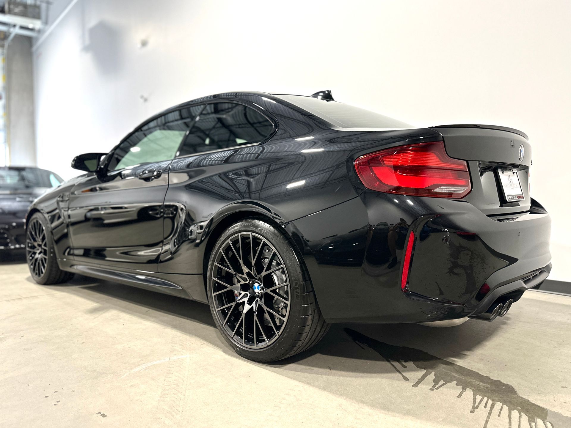 Black BMW M2 coupe, rear view, parked inside. Shiny paint, black rims, red tail lights.