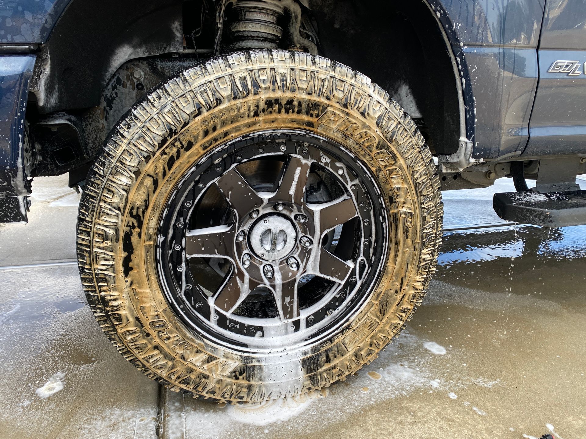 Truck tire being washed, covered in soap and mud. Black rim, side of blue truck.
