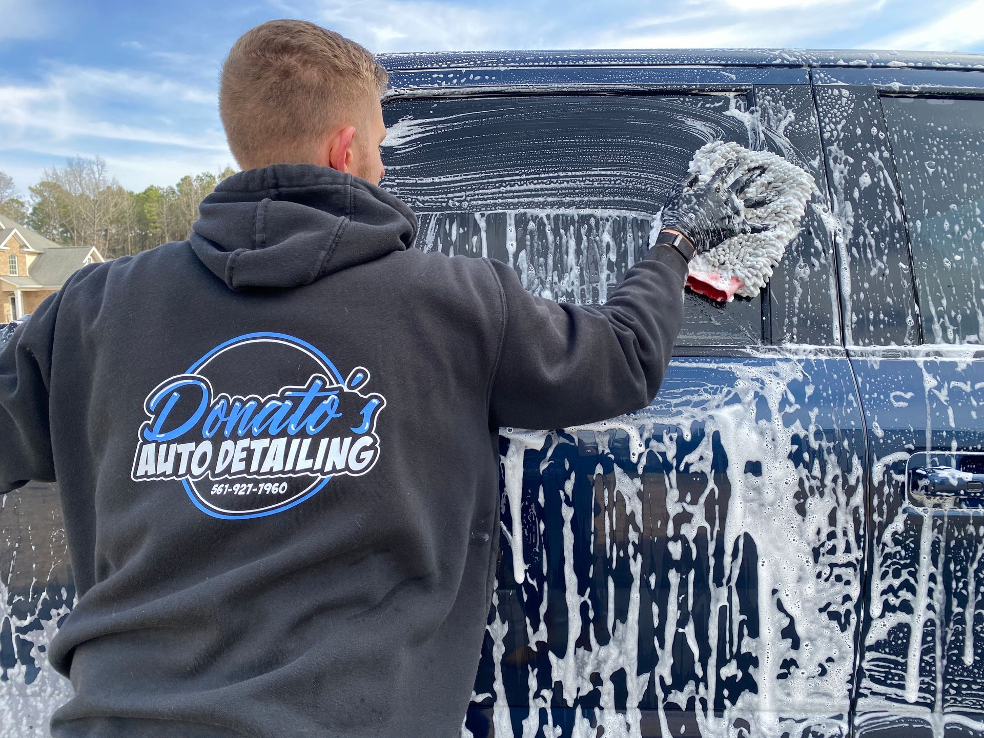 Man washing a blue car with soapy water, wearing a hoodie with 