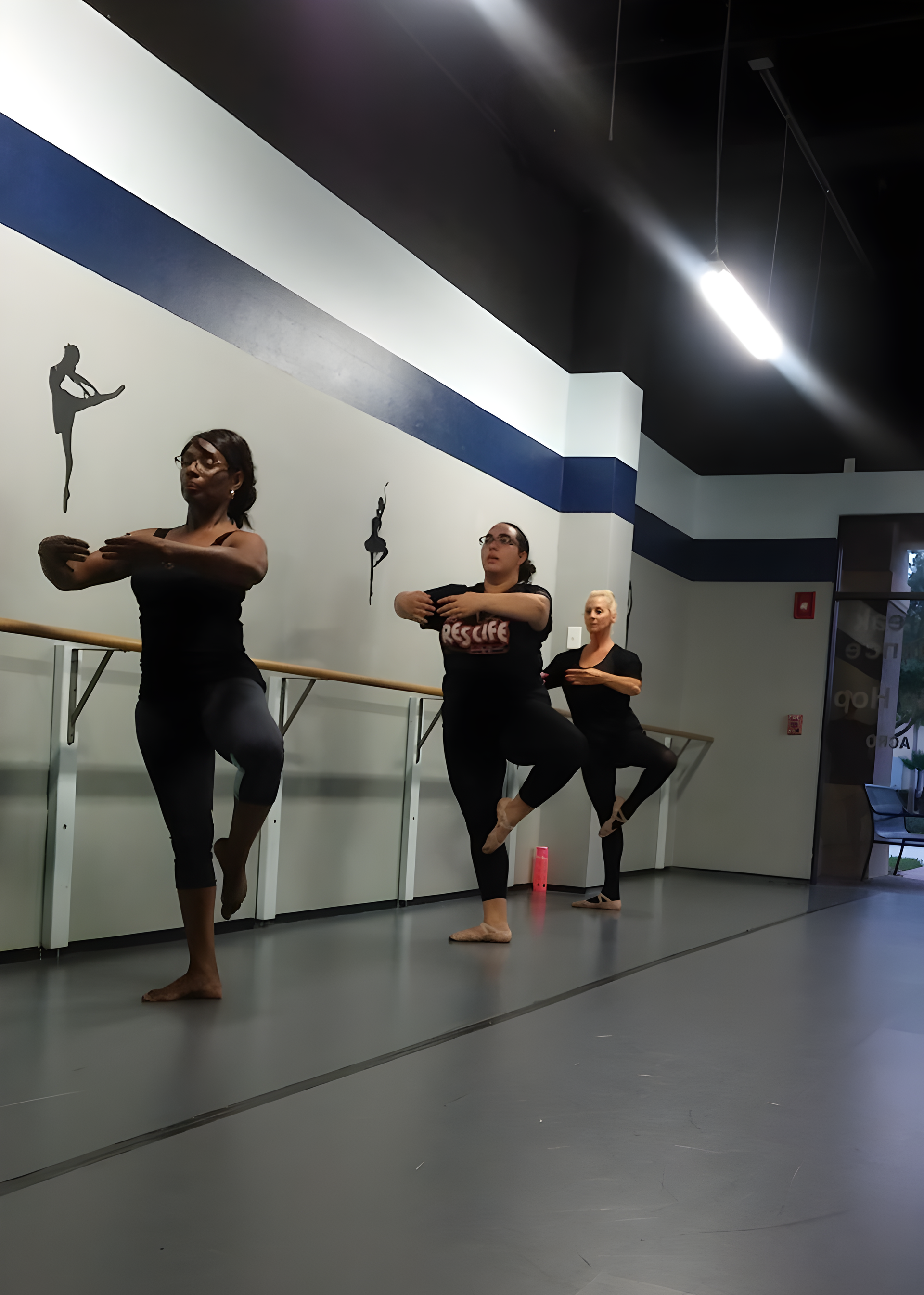 Three women practice ballet at a barre in a dance studio. They are in a pose with one leg raised.