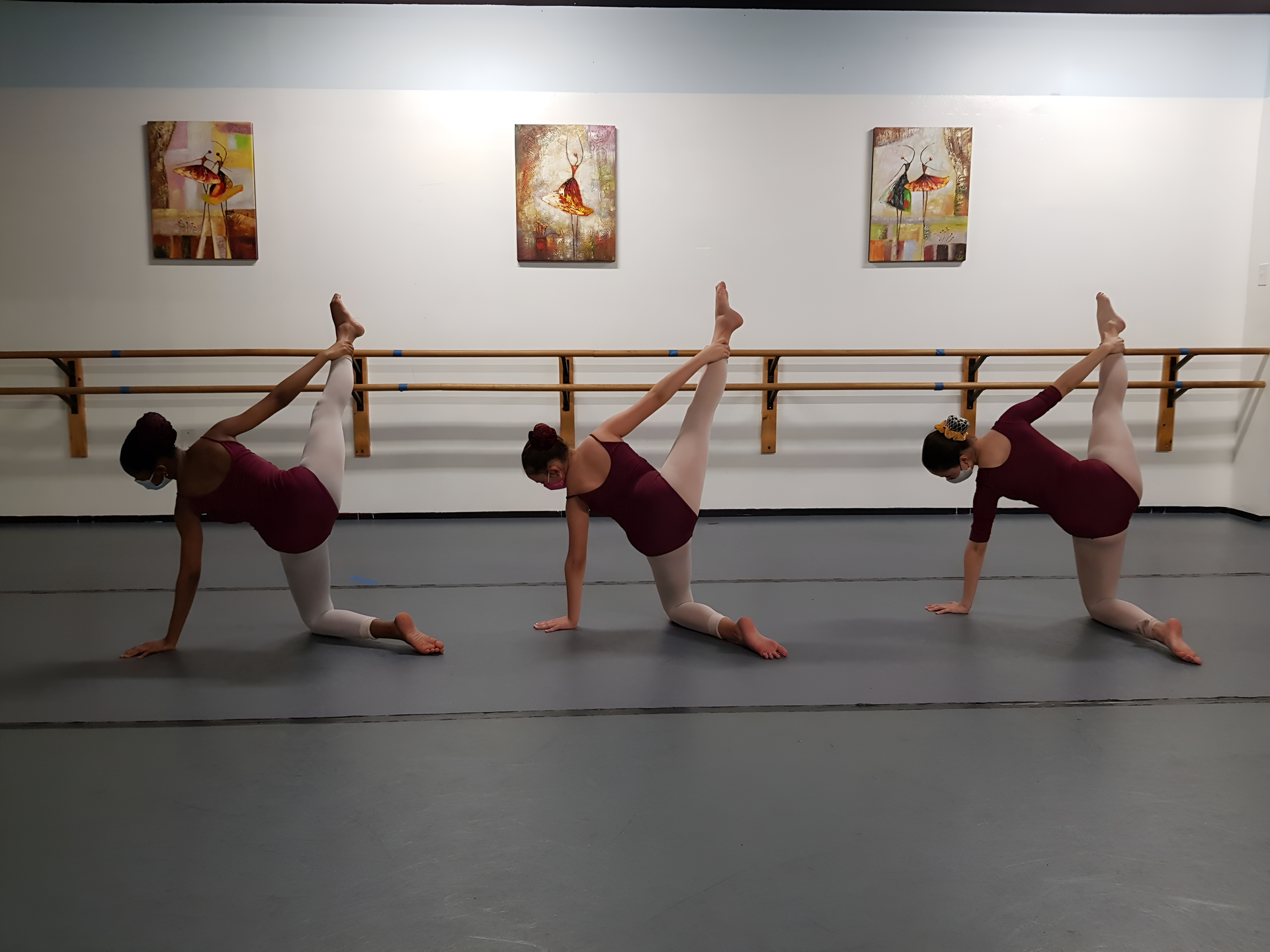 Three ballet dancers in a studio, each in a kneeling arabesque, burgundy tops, and light pants.
