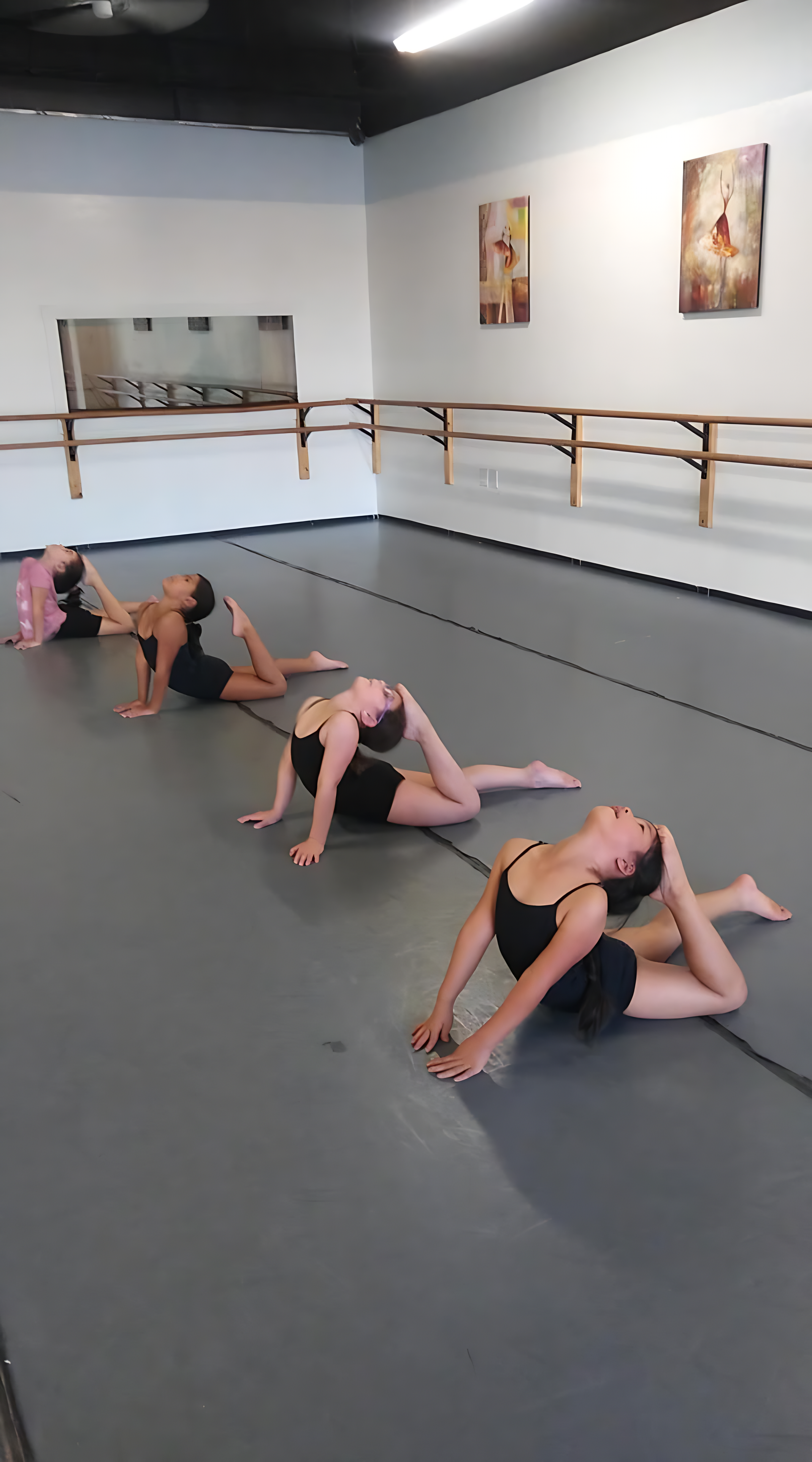 Dancers stretching on a gray floor in a studio, arms arched back. They are wearing black leotards.