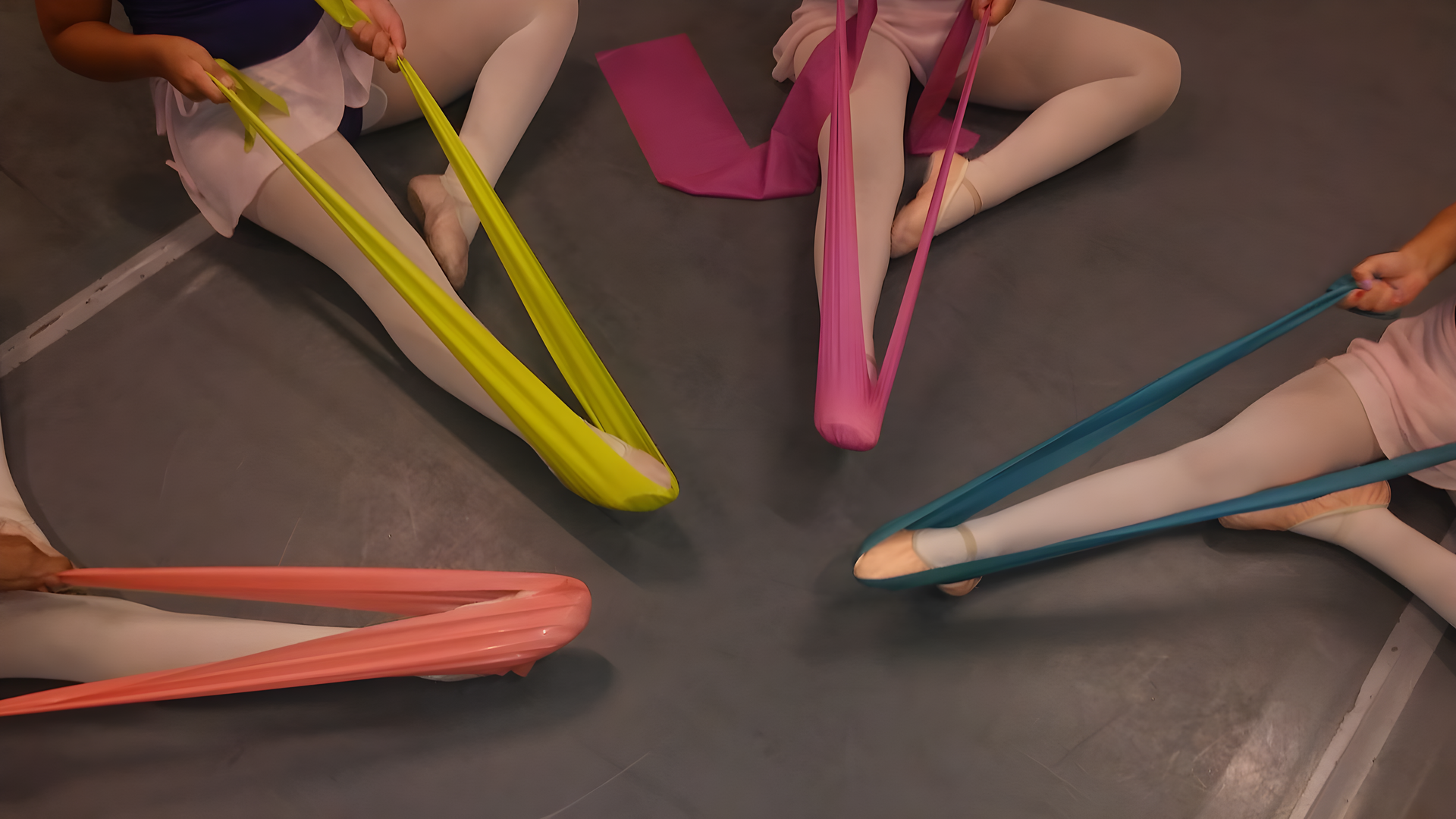 Dancers seated, stretching feet with colorful resistance bands in a dance studio.