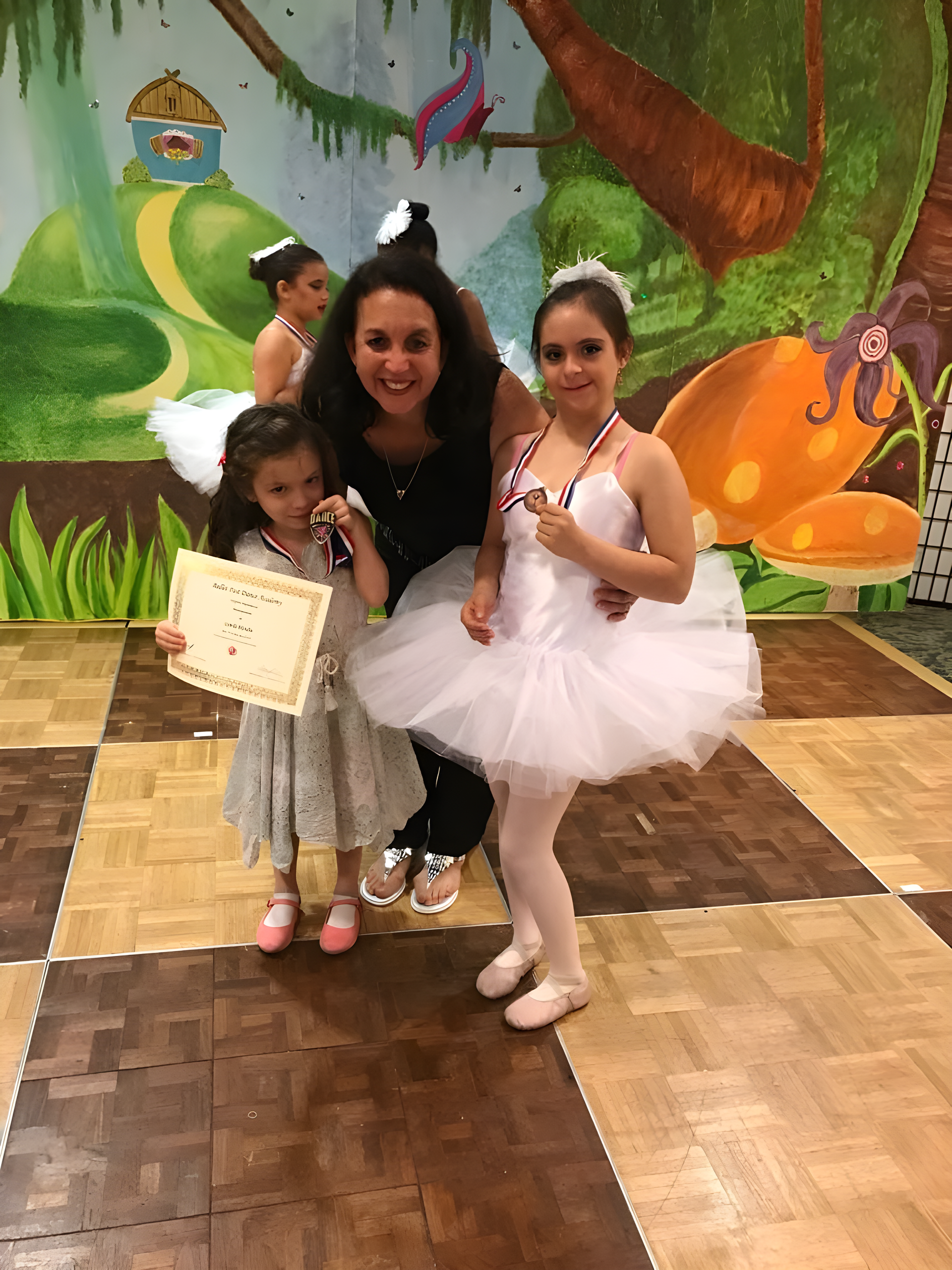 Woman with two young girls in ballet attire, holding awards, smiling in a studio.