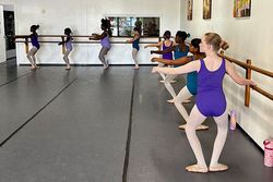 Young dancers in colorful leotards practice ballet at a barre in a studio with wooden accents.