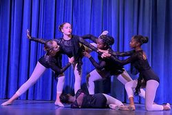 Dancers in black leotards and white leggings perform a modern dance routine on a stage with blue curtains.