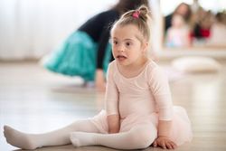 A young girl with Down syndrome in ballet attire sits on the floor, looking to her side.
