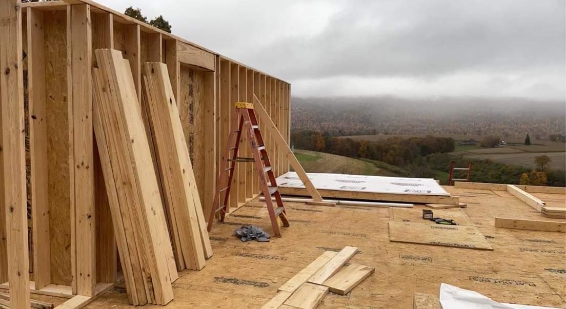 Construction of a wooden house frame on a cloudy day with a view of a forested landscape.