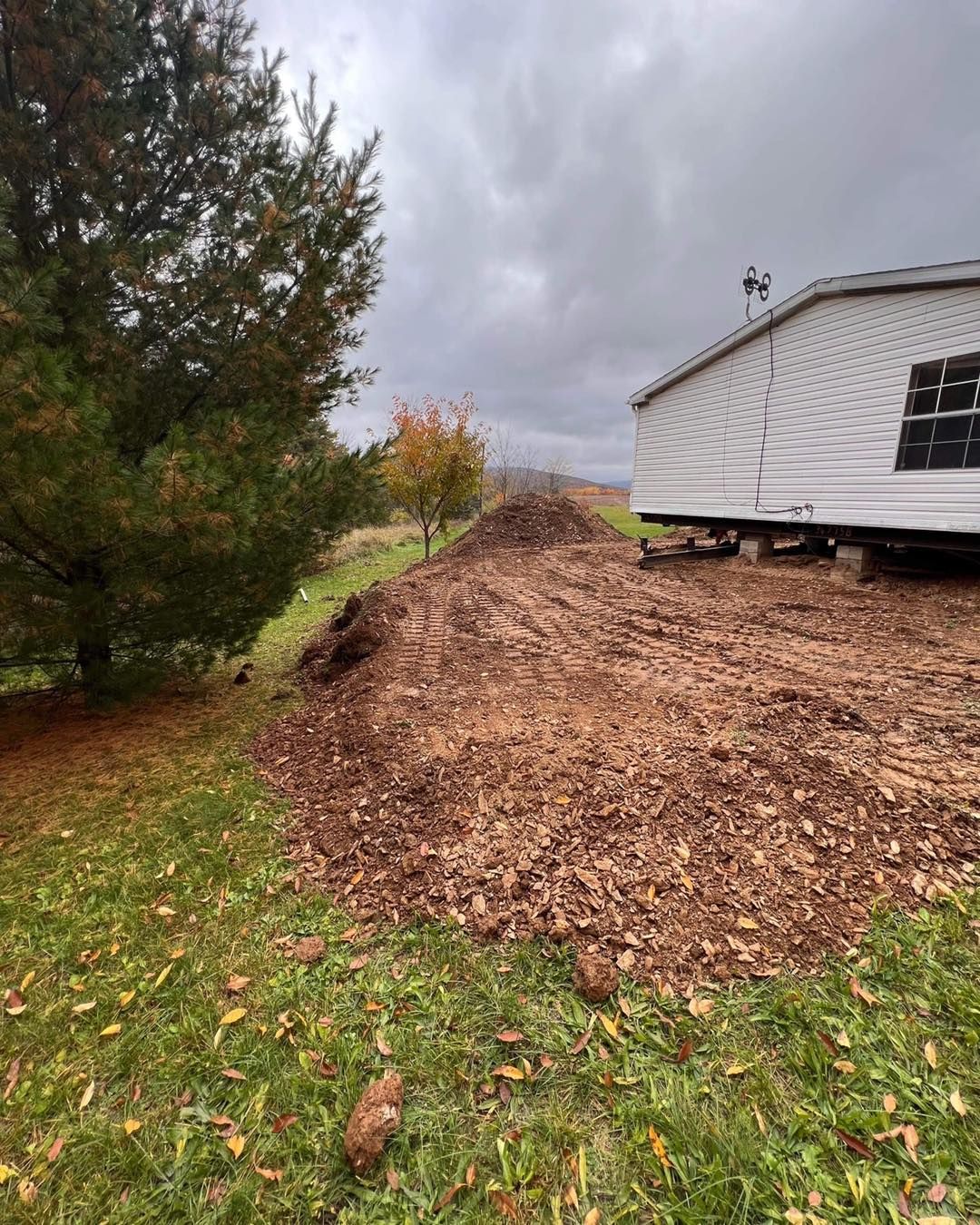 Large pile of brown mulch next to a mobile home and green lawn under a cloudy sky.