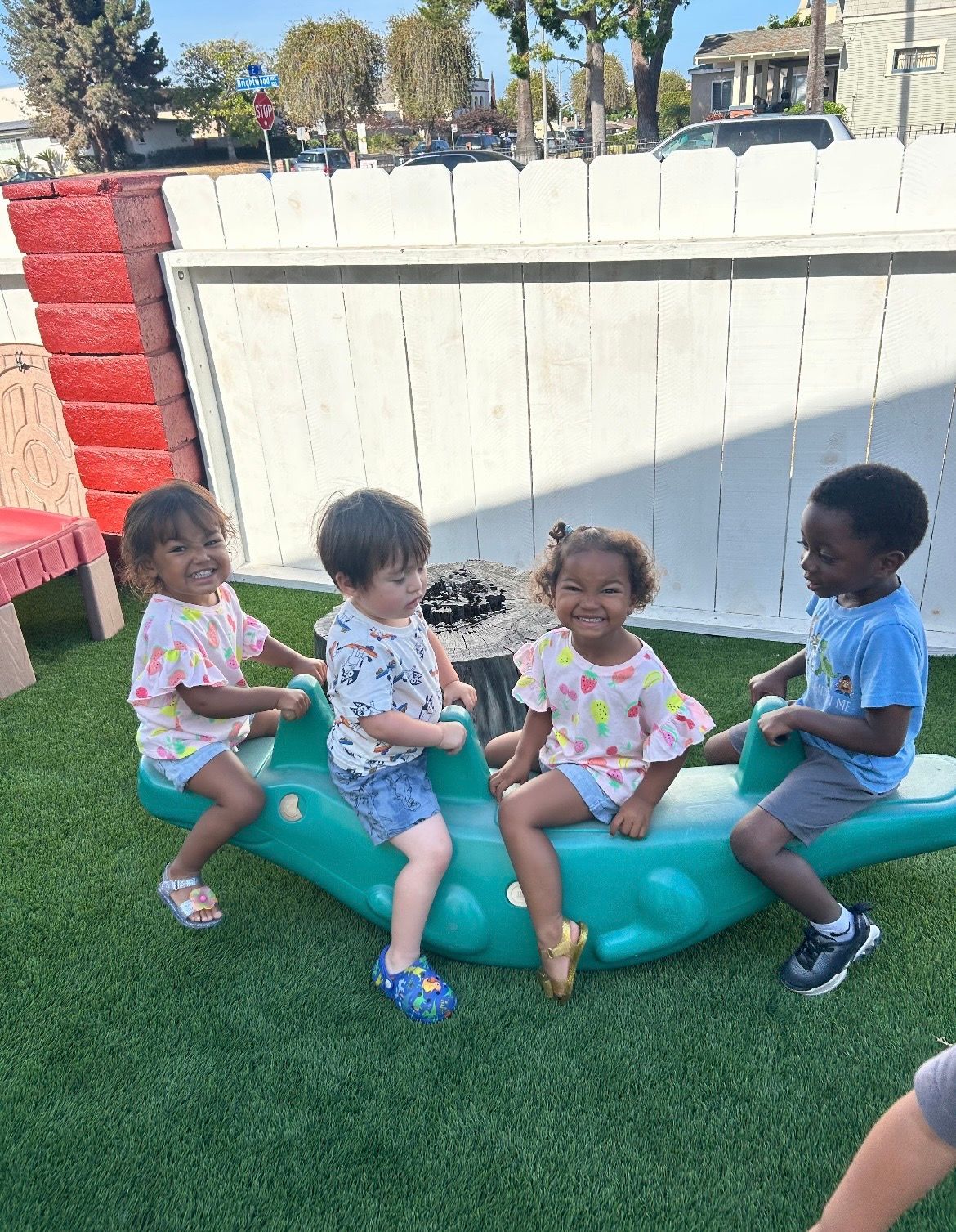 Four children sit on a turquoise teeter-totter in a grassy backyard, smiling in the sun.